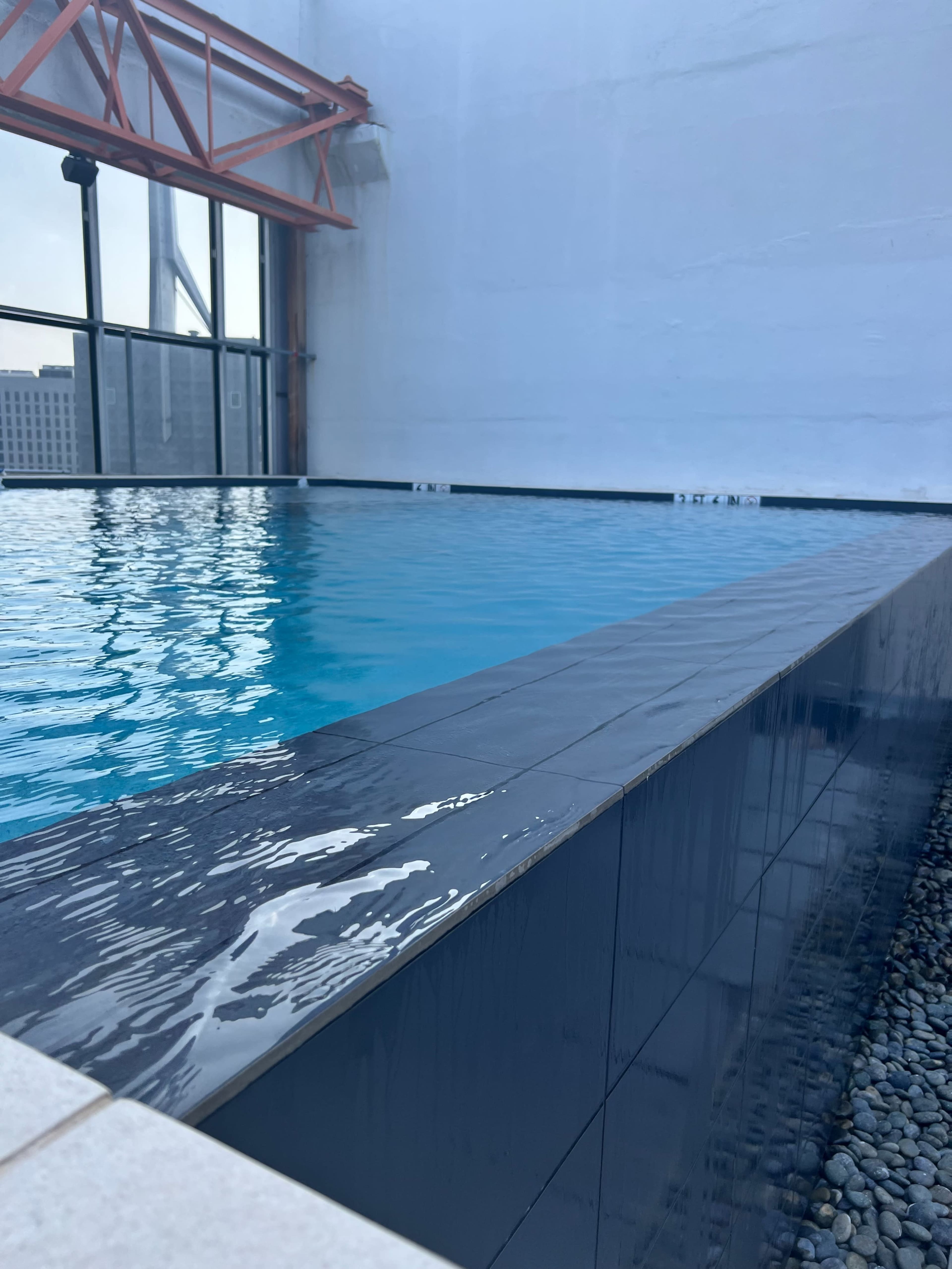 A rooftop swimming pool with a sleek dark edge reflecting the water's surface, situated next to a glass wall.