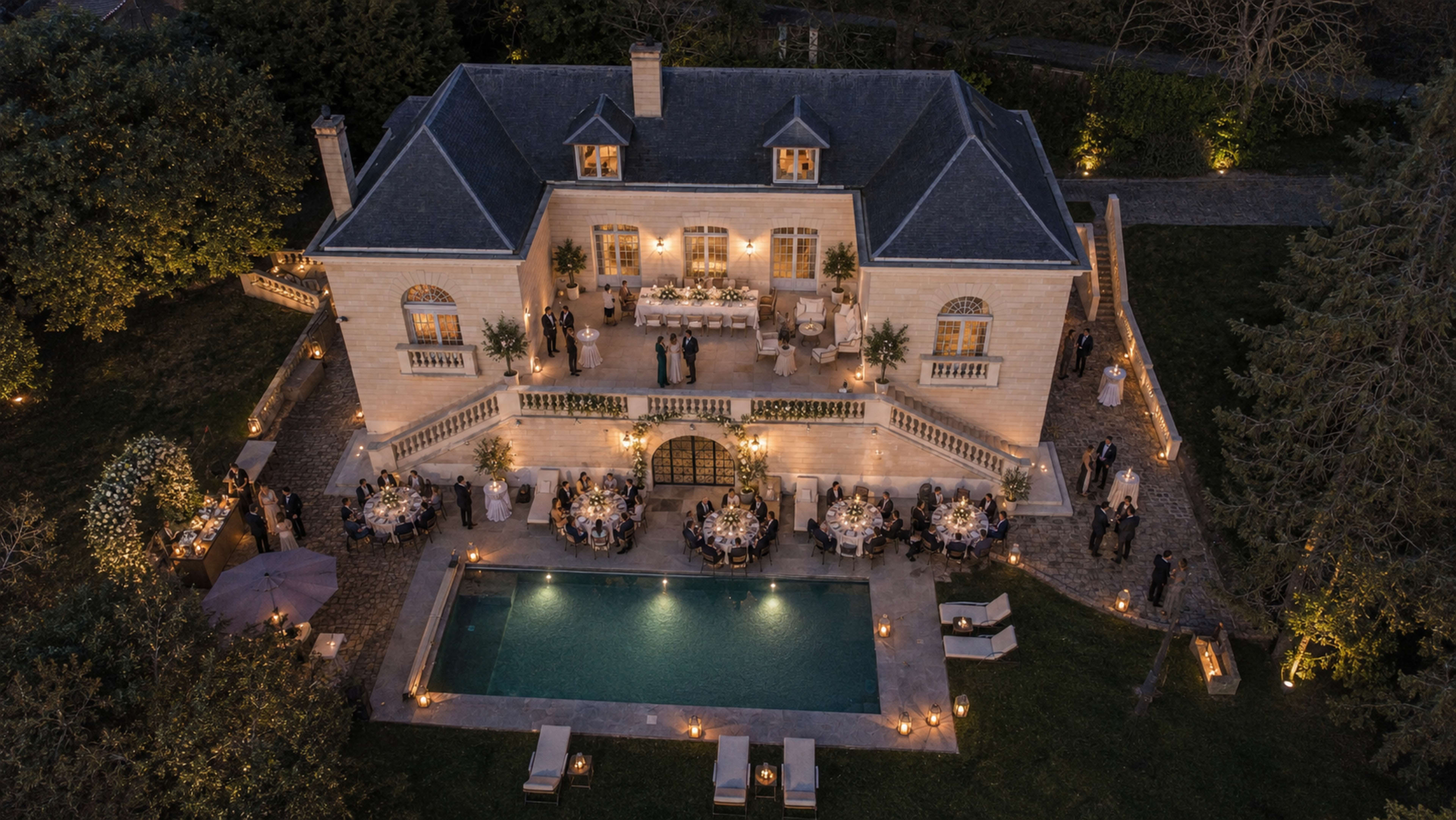 An elegant mansion with a pool is hosting an outdoor event, surrounded by guests and illuminated by decorative lighting.