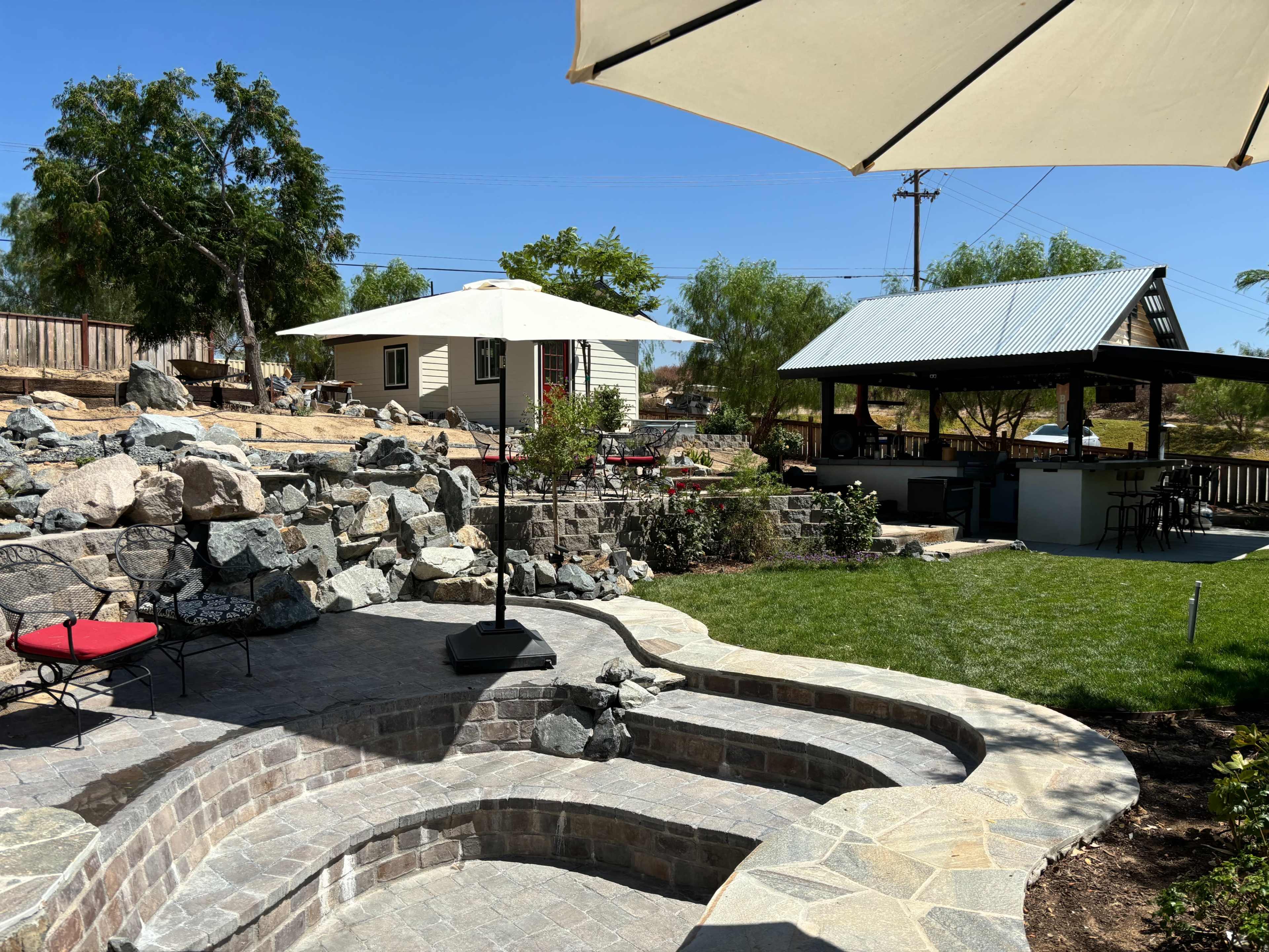 A patio area with stone steps, a red chair, and an umbrella, surrounded by a grassy lawn, a rock garden, and a small building in the background.