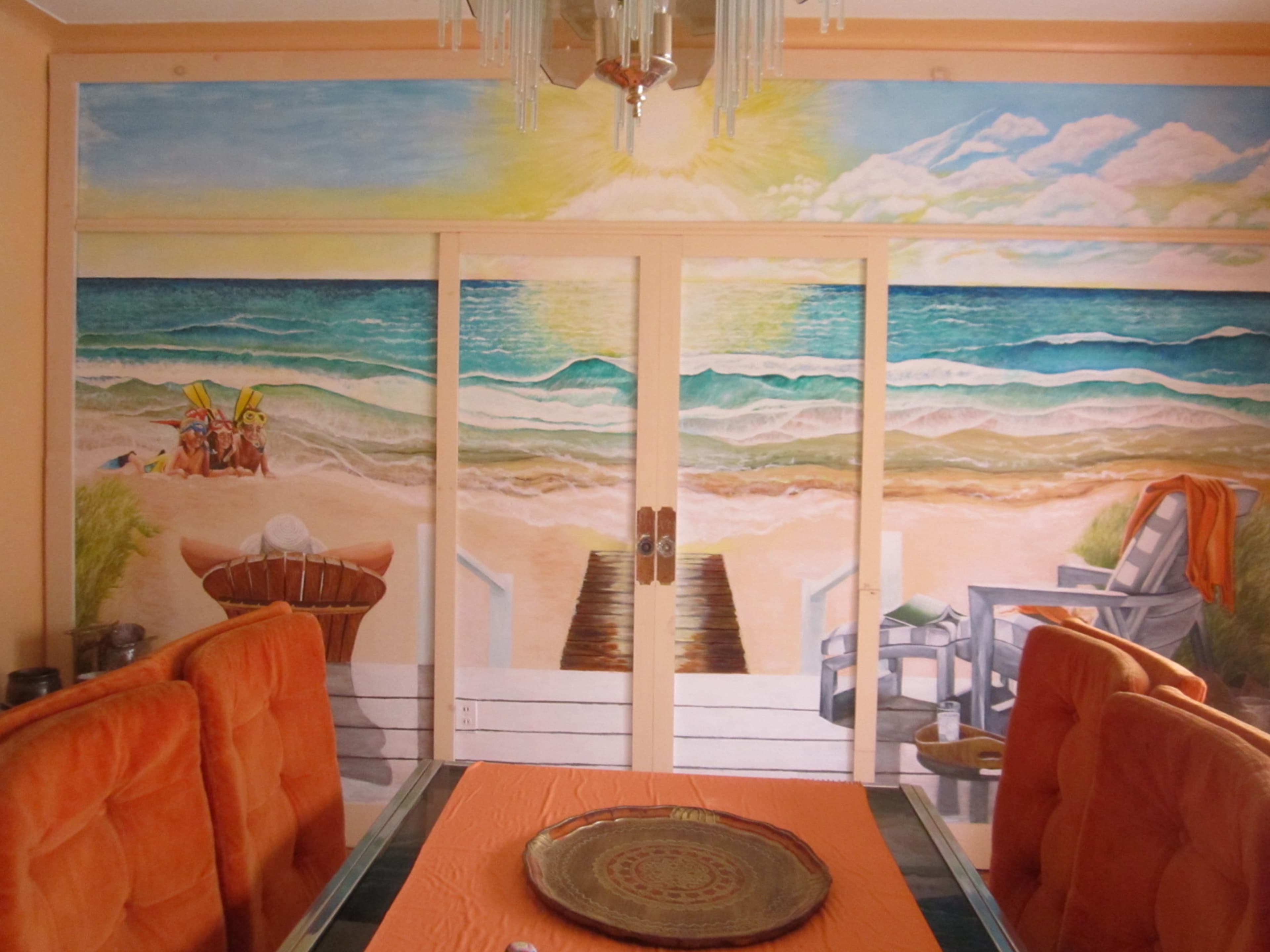 A dining area with an orange tablecloth and upholstered chairs, complemented by a large mural depicting a beach scene with waves, a sandy shore, and sunlit skies.