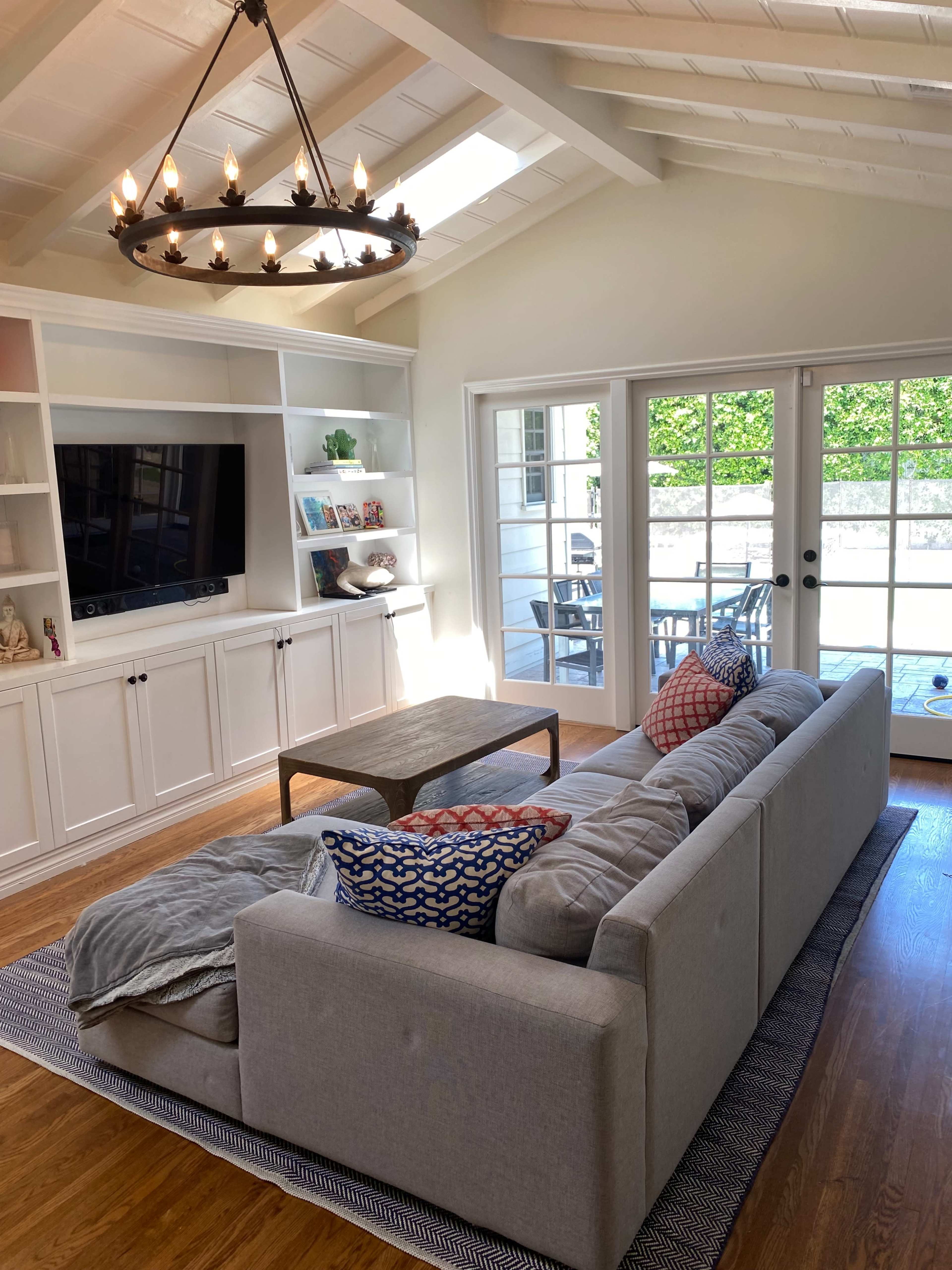 A modern living room features a light-colored sectional sofa with patterned pillows, a coffee table, and built-in shelves, with large glass doors leading to an outdoor patio.