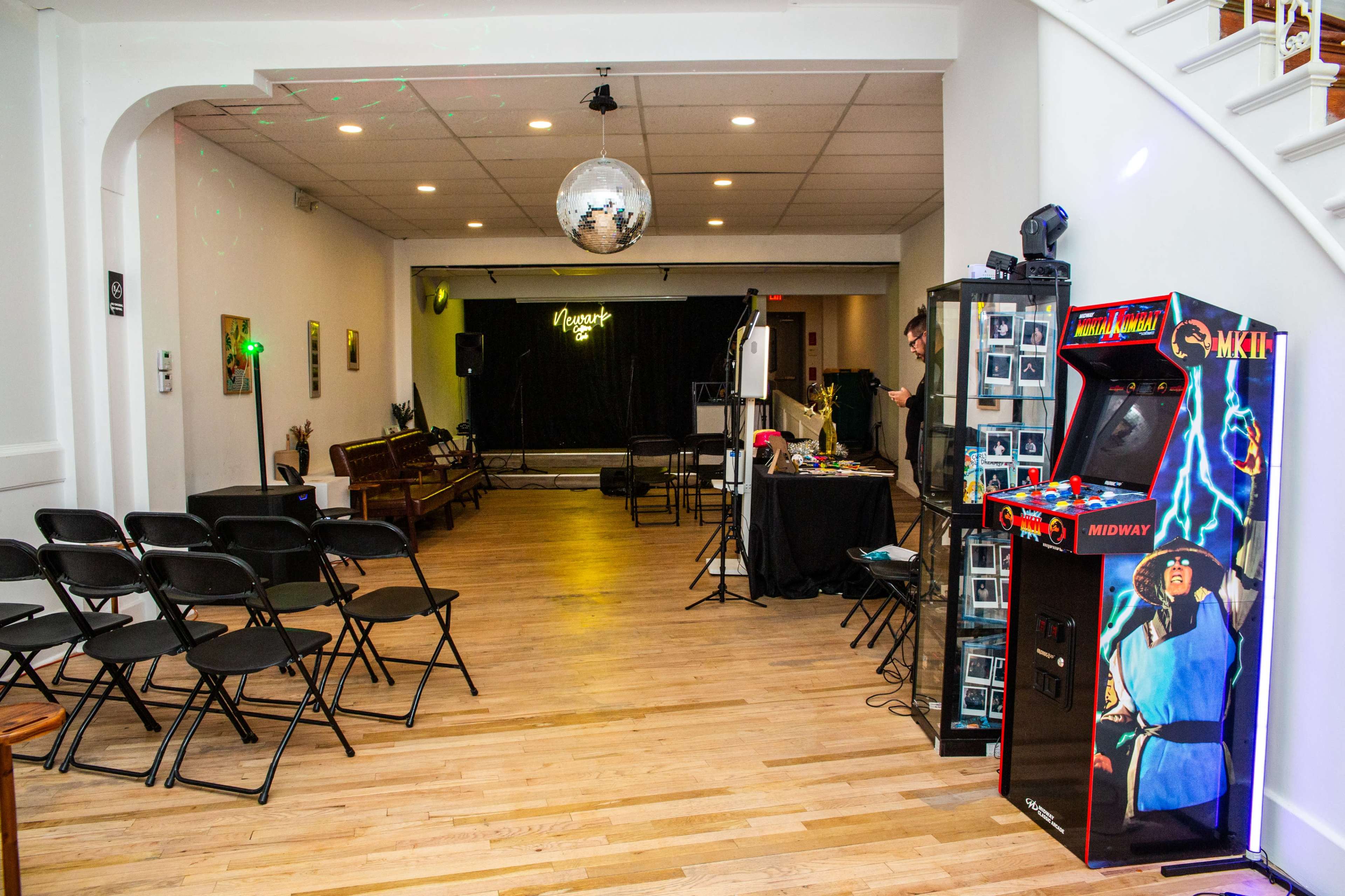 The image shows a spacious room set up for an event, featuring a stage area, folding chairs arranged for seating, and a vintage arcade machine on one side.