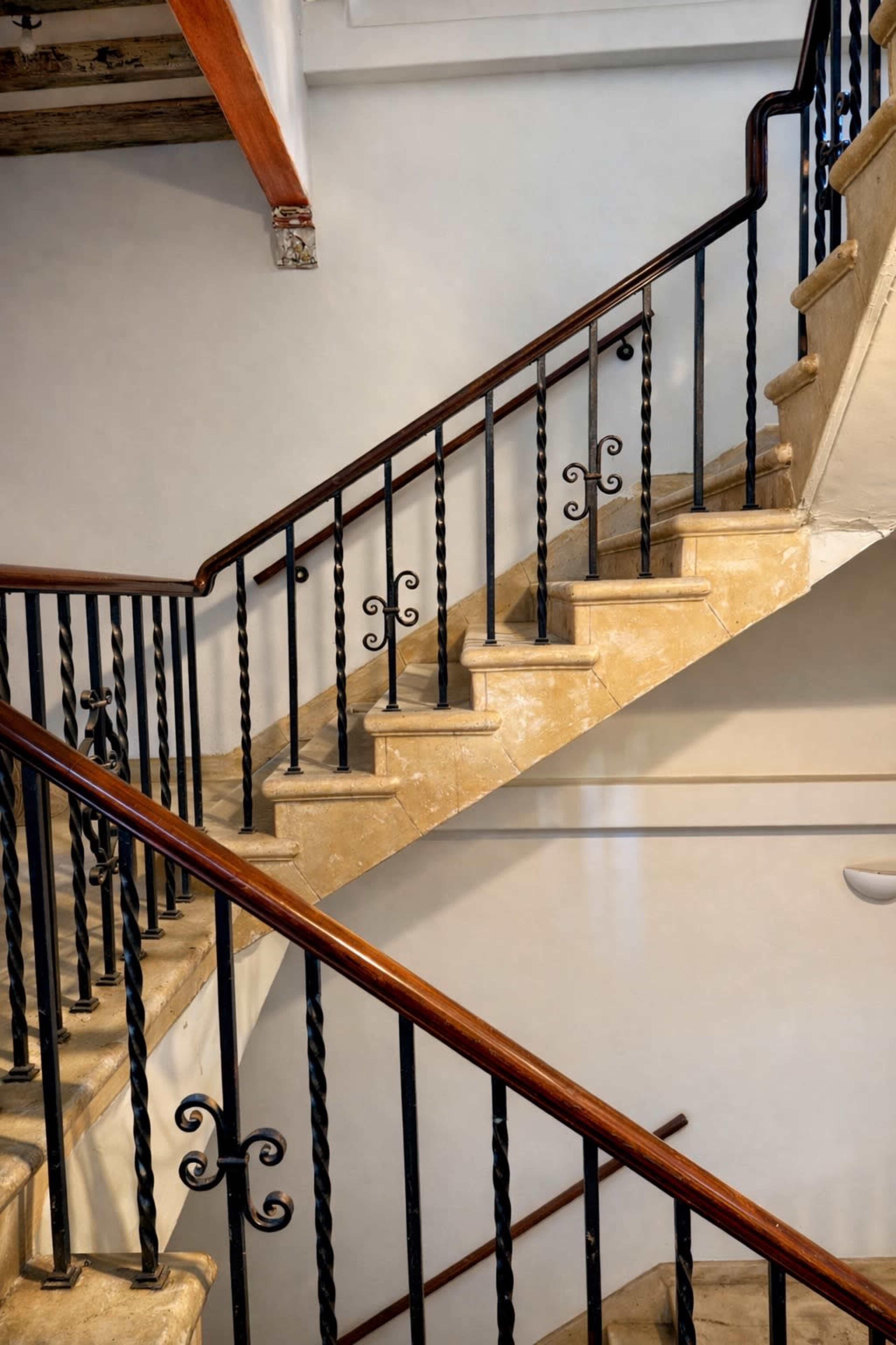 The image shows a stairwell with a wrought iron railing and a stone staircase, featuring two landings and a smooth, light-colored wall.