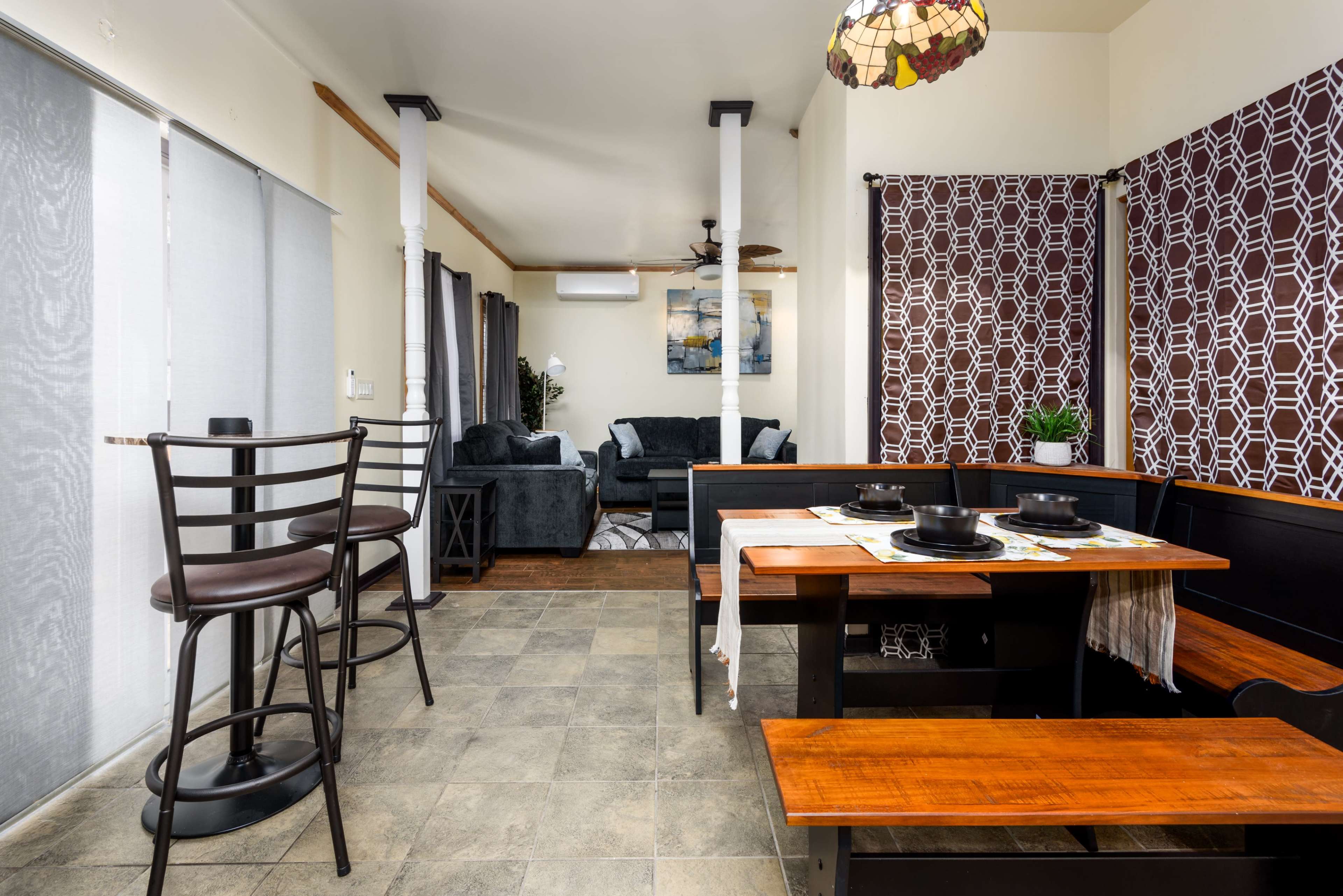 A dining area with a wooden table and benches, bar stools, and a living room visible in the background with a blue couch and decorative accents.