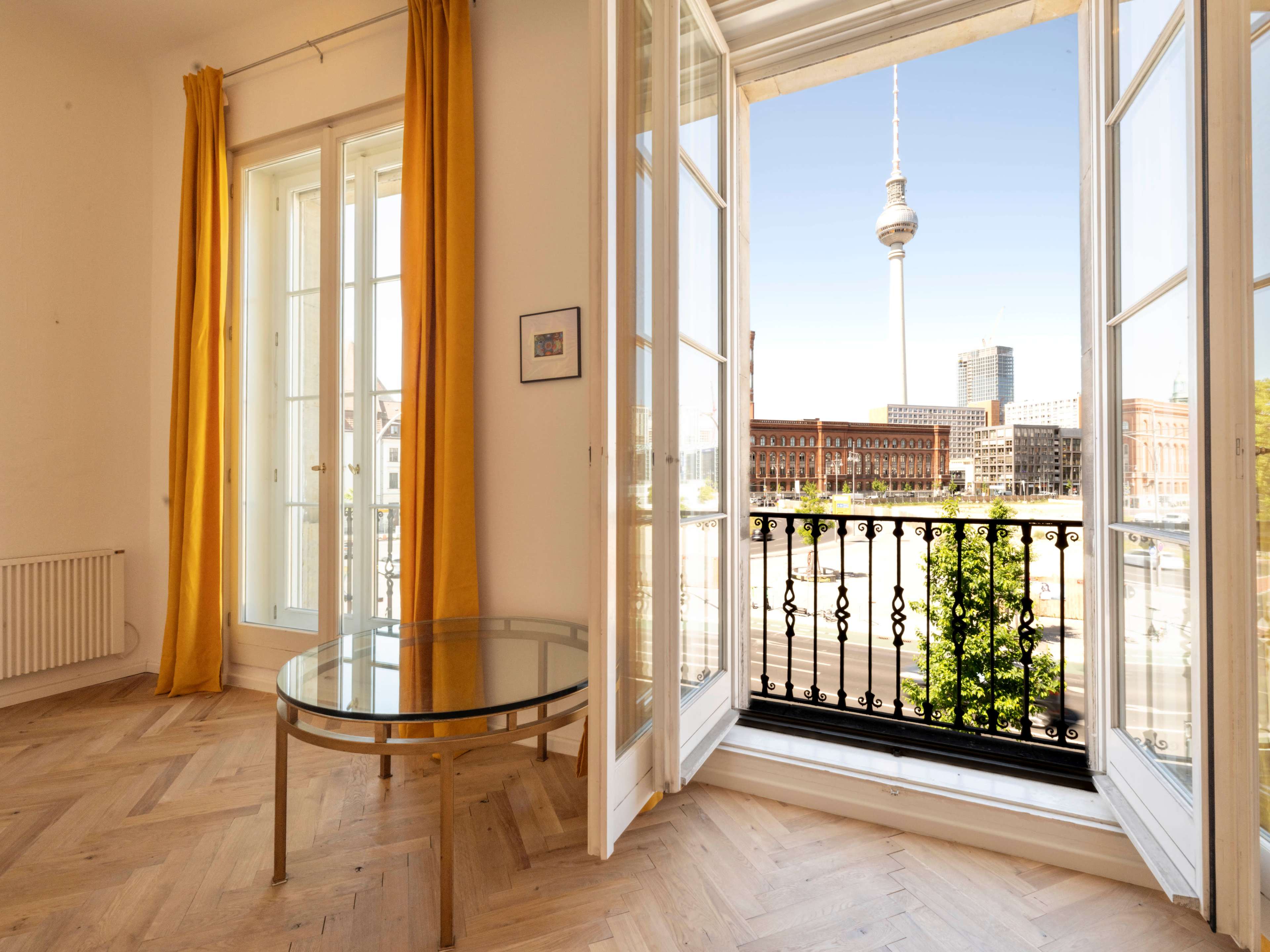 A bright room with yellow curtains features a glass table and open French doors that overlook the Berlin TV Tower and urban landscape.