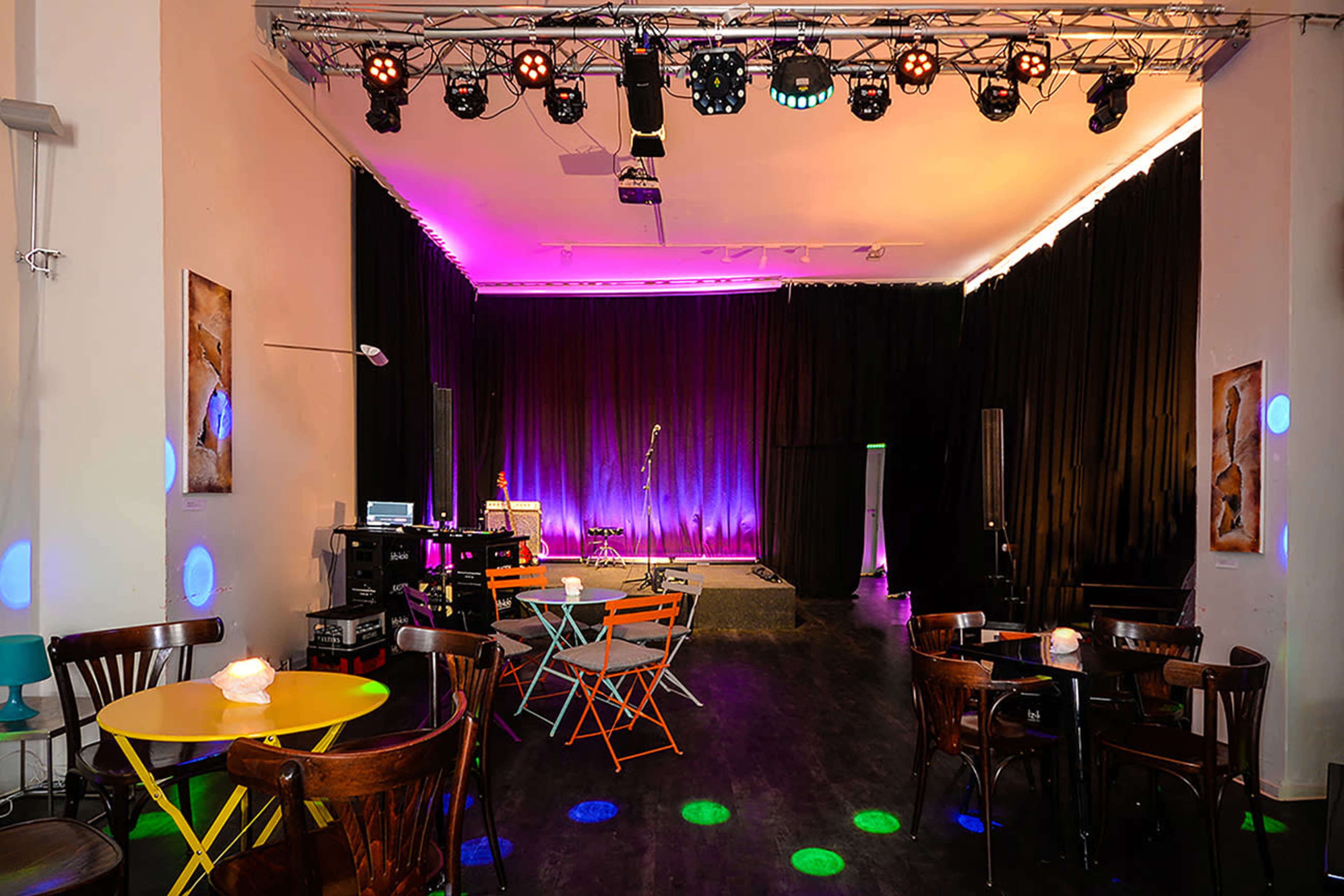 A small stage area in a performance venue with colorful lighting, surrounded by tables and chairs.