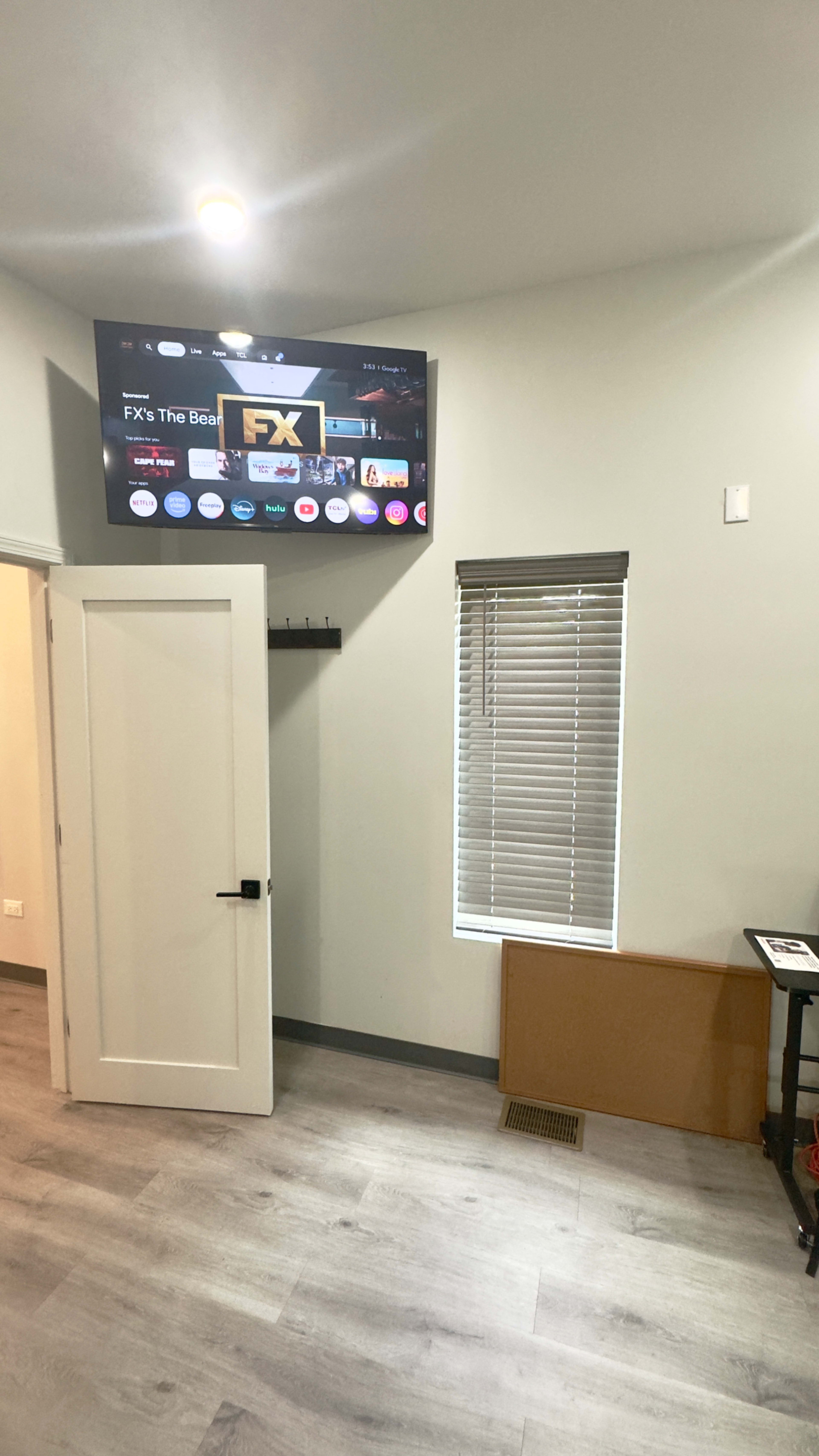 The image shows a room with a flat-screen TV mounted on the wall, a door leading to another space, a window with blinds, and a small table or shelf in the corner.