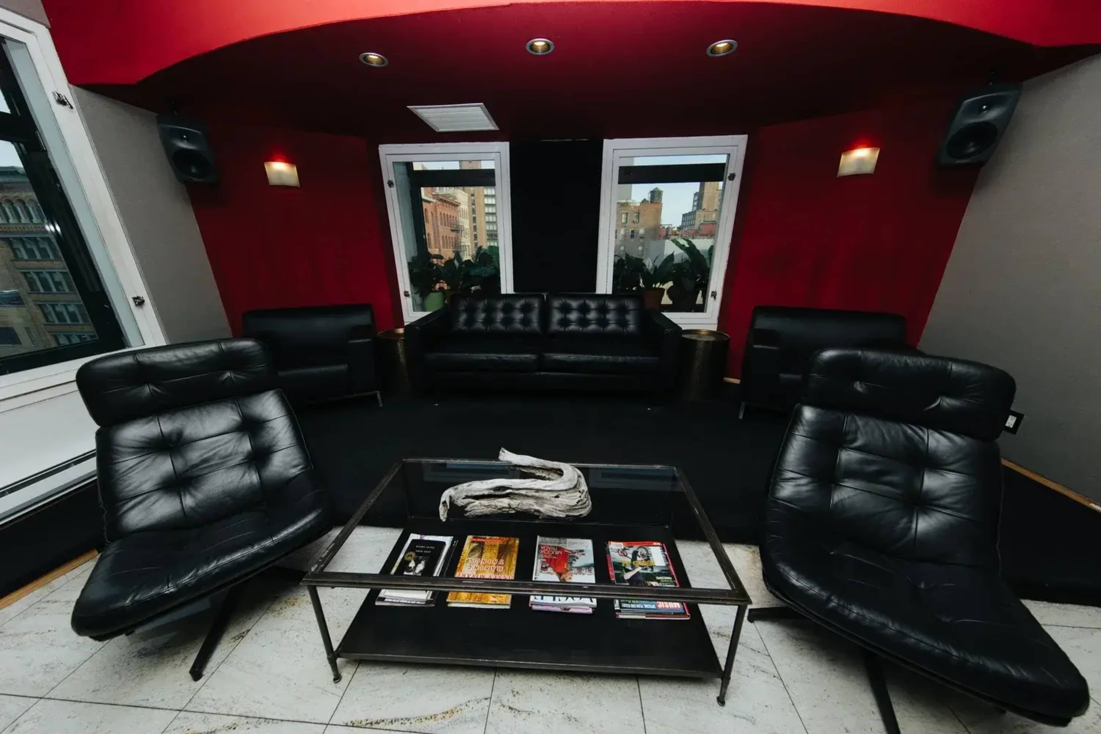 The image shows a modern lounge area with black leather furniture, including a sofa and two armchairs, arranged around a glass coffee table, against a backdrop of red and gray walls.