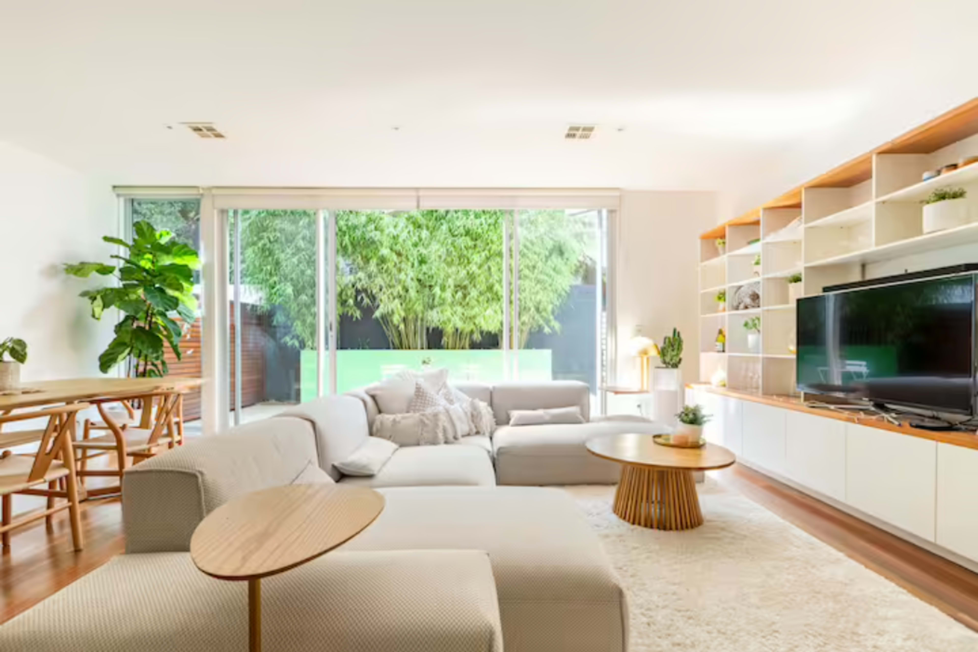 A modern living room features a sectional sofa, a round coffee table, and a large window overlooking a green outdoor space.