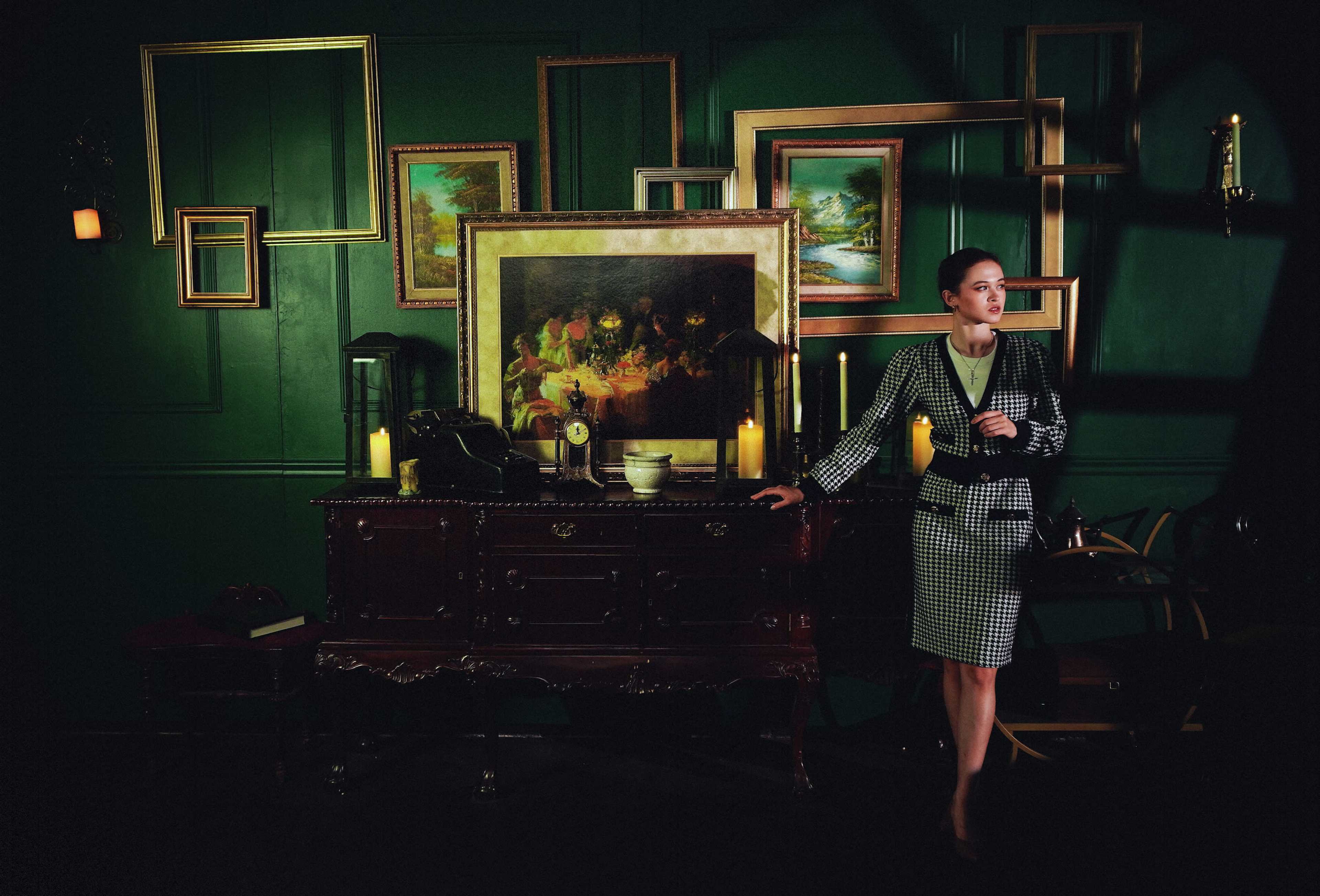 A woman stands beside a dark wooden cabinet adorned with candles and framed paintings in a dimly lit room.