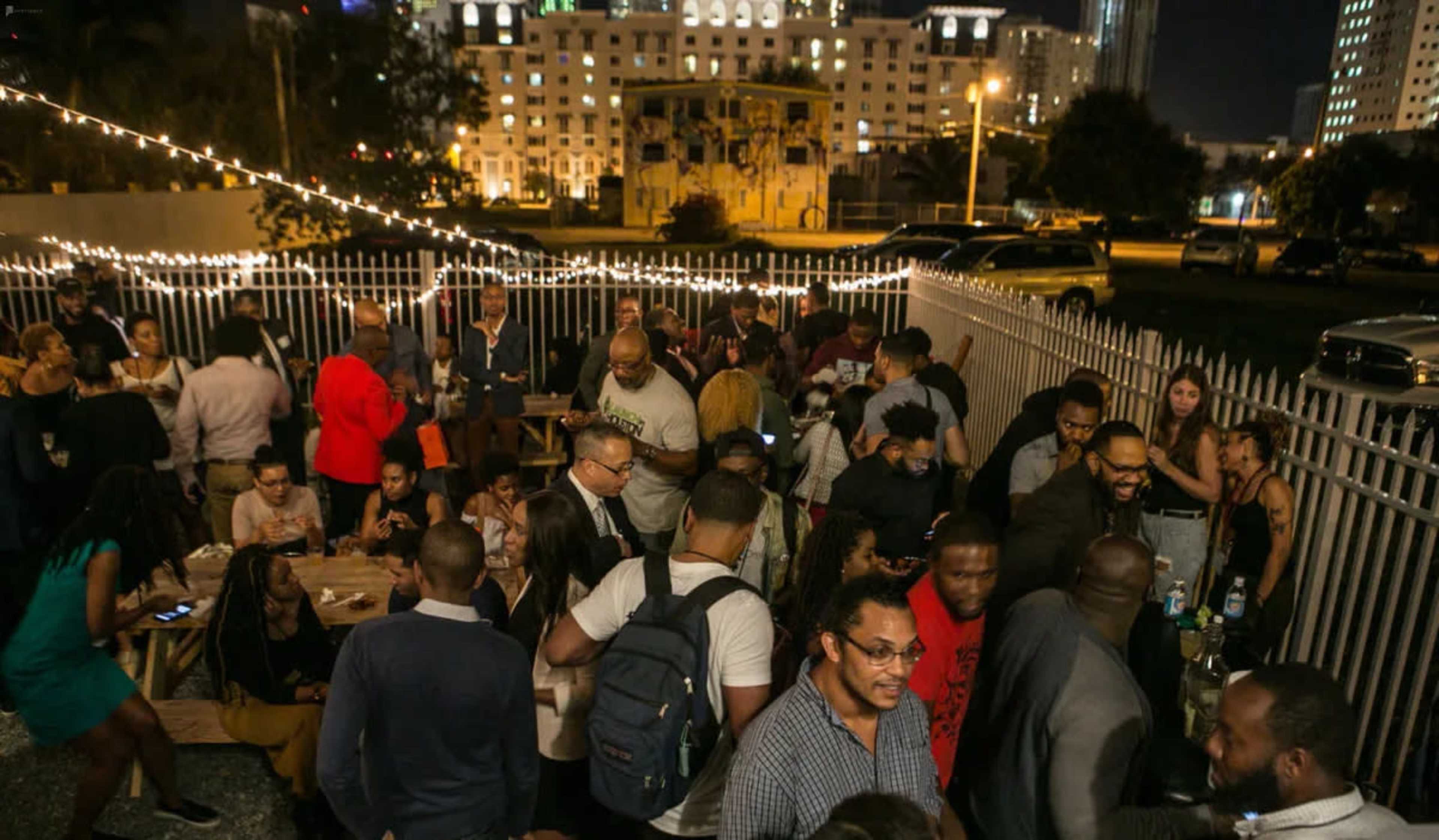 A crowded outdoor gathering takes place at night, featuring people socializing and mingling around tables illuminated by string lights.