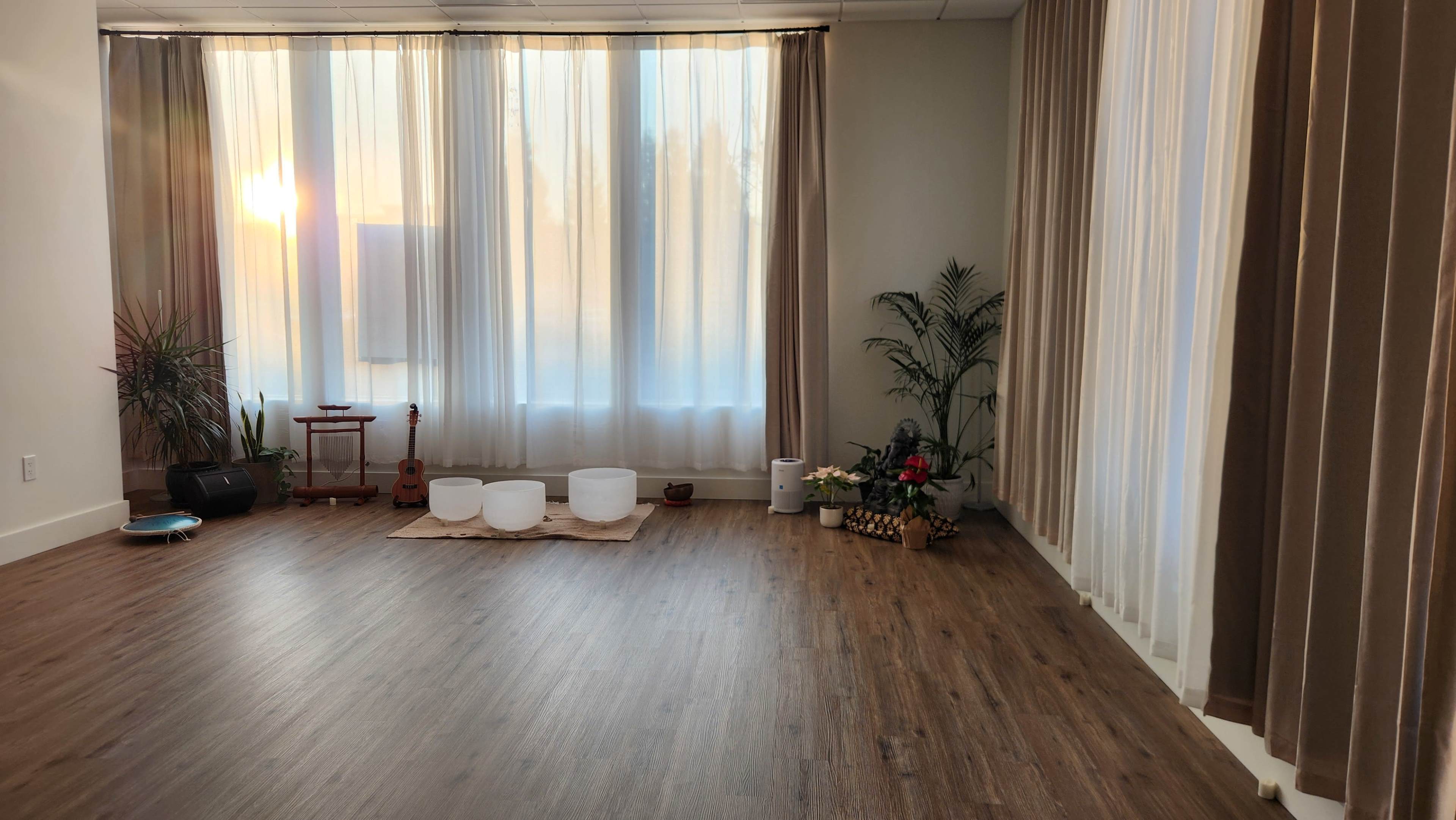 The image shows a serene room with large curtains allowing sunlight to filter in, featuring a wooden floor, two crystal singing bowls on a mat, and a potted plant.