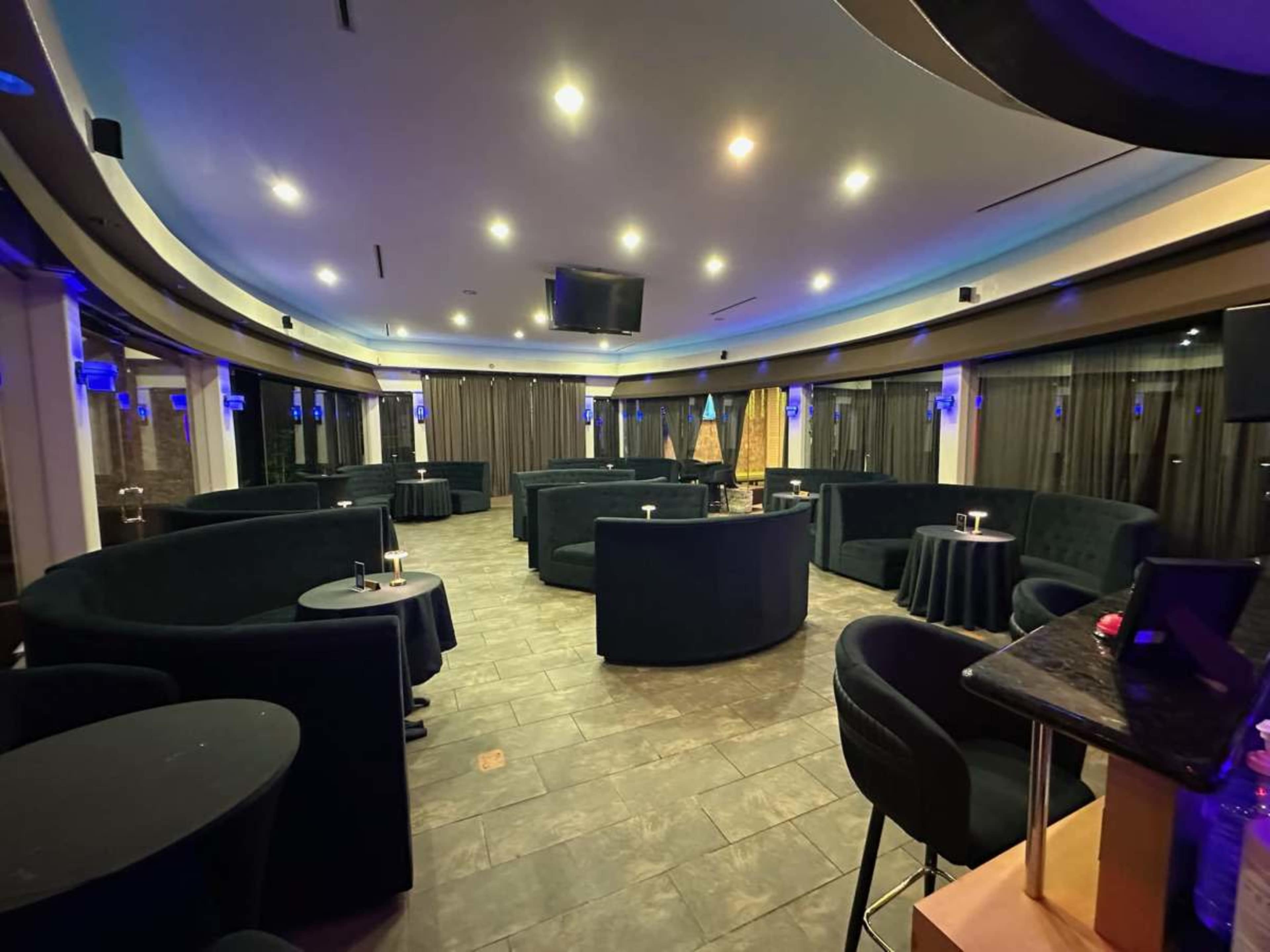 The image shows a dimly lit lounge with circular seating arrangements and small tables, featuring a contemporary design and blue accent lighting.