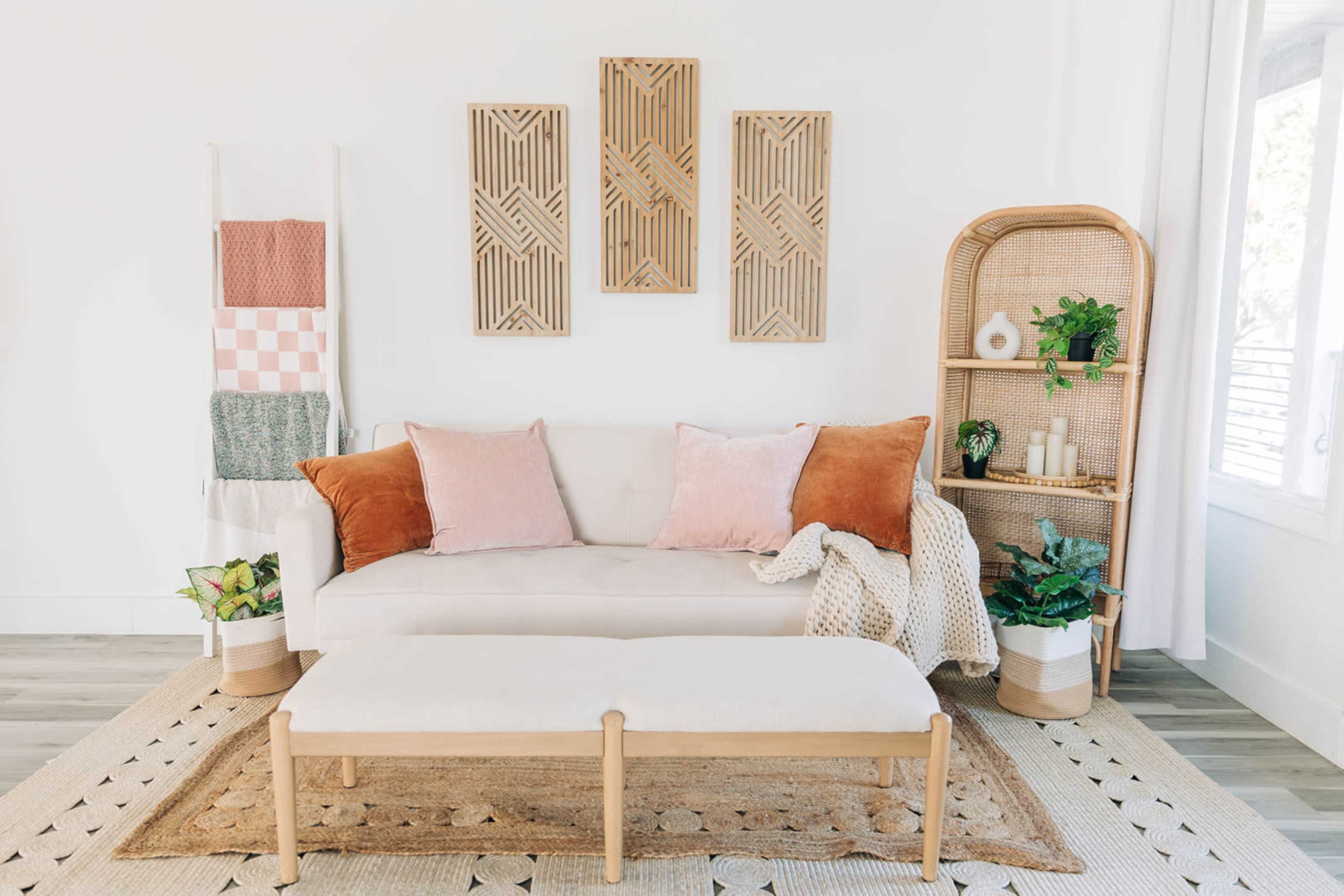 A modern living room features a light couch with assorted pillows, a wooden bench, and decorative wall art, alongside plants and a woven shelf.