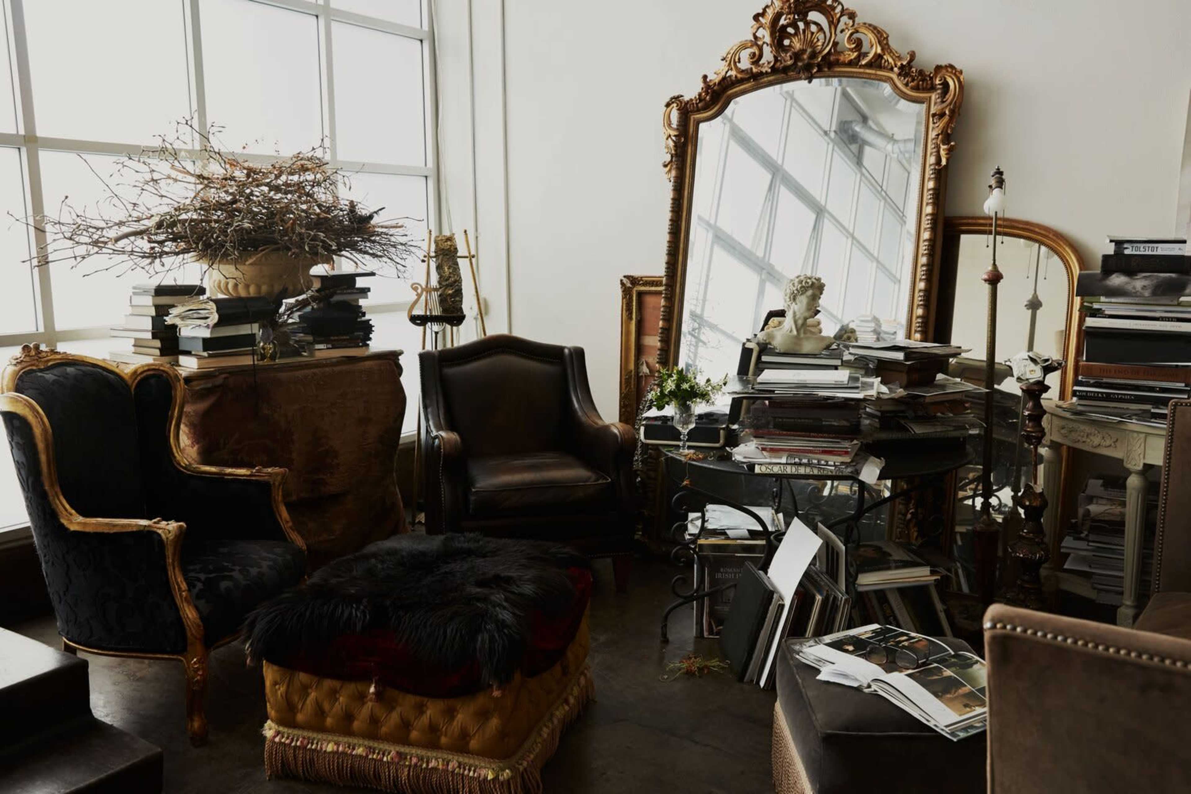 The scene features a stylishly cluttered room with a vintage mirror, ornate furniture, and stacks of books and magazines on a glass table.