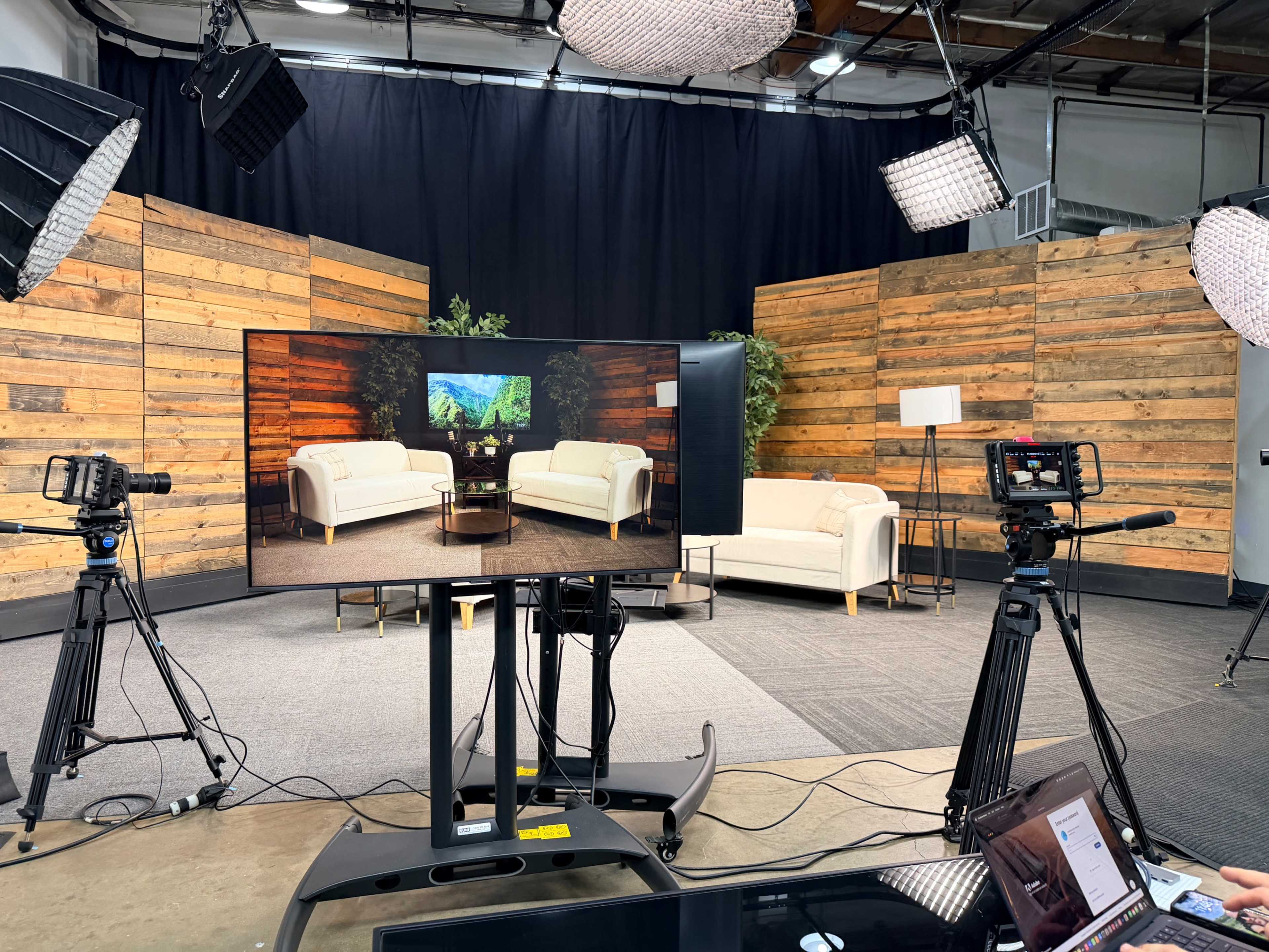 The image shows a studio setup for filming, featuring two white sofas, a coffee table, and wooden backdrop panels, with cameras and lighting equipment in place.