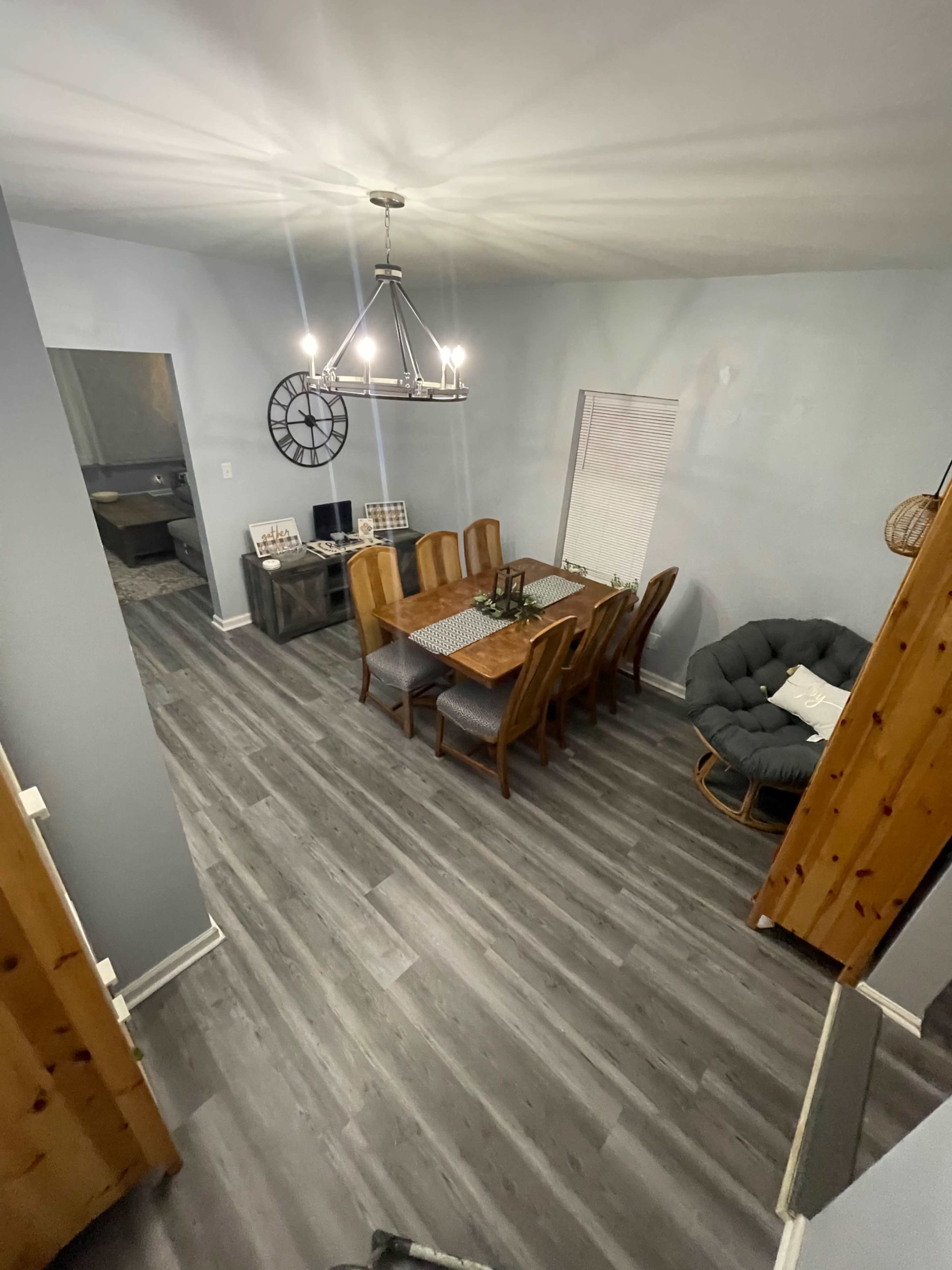 A dining area with a long wooden table surrounded by chairs, a chandelier above, and gray walls and flooring.