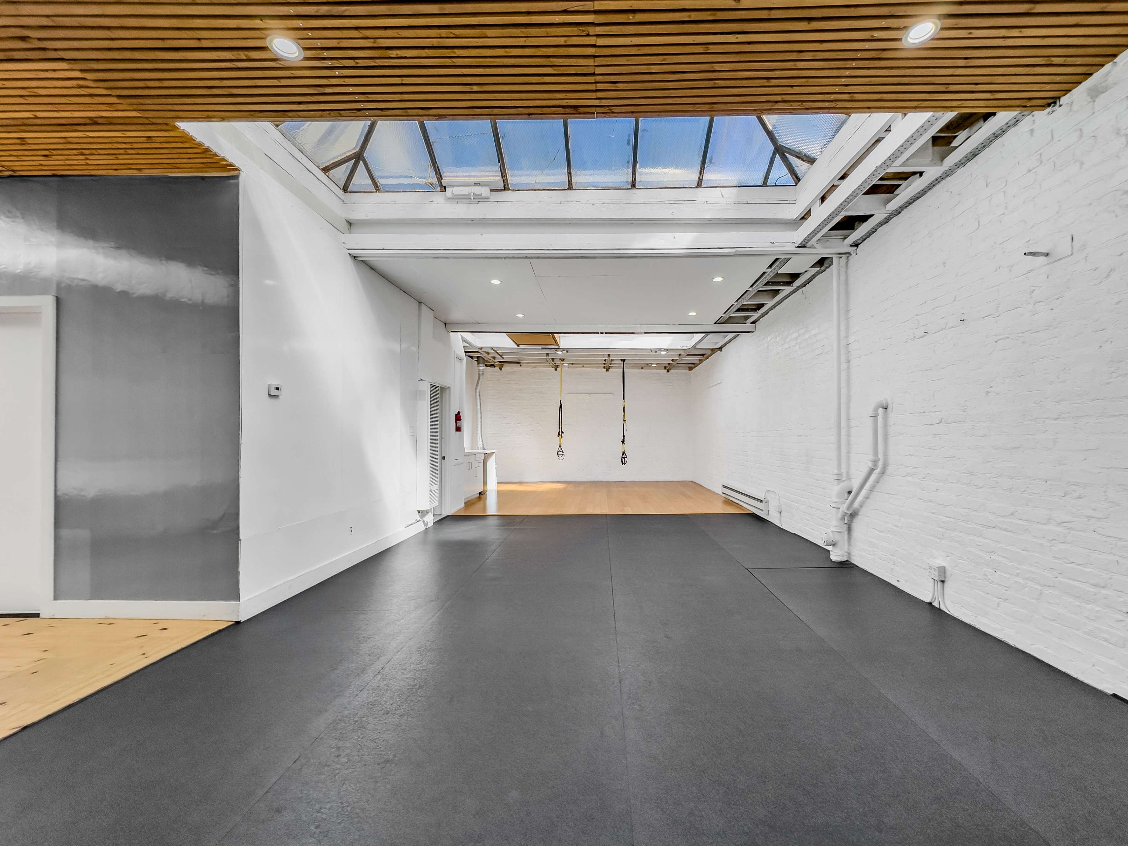 A spacious, well-lit room with a black rubber floor, white walls, and high wooden ceilings, featuring exercise equipment hanging from the ceiling.