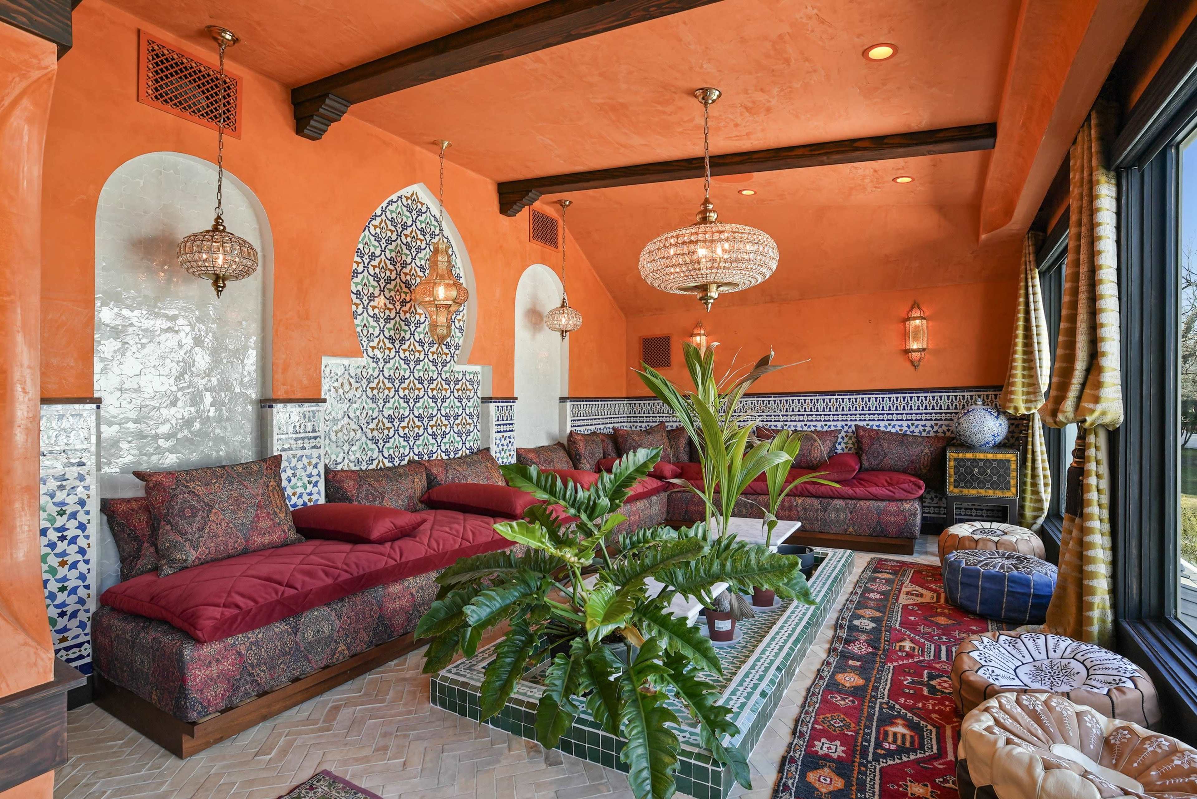 The image shows a richly decorated room with orange walls, comfortable seating adorned with red cushions, and intricate tile work, featuring hanging light fixtures and a potted plant in the center.