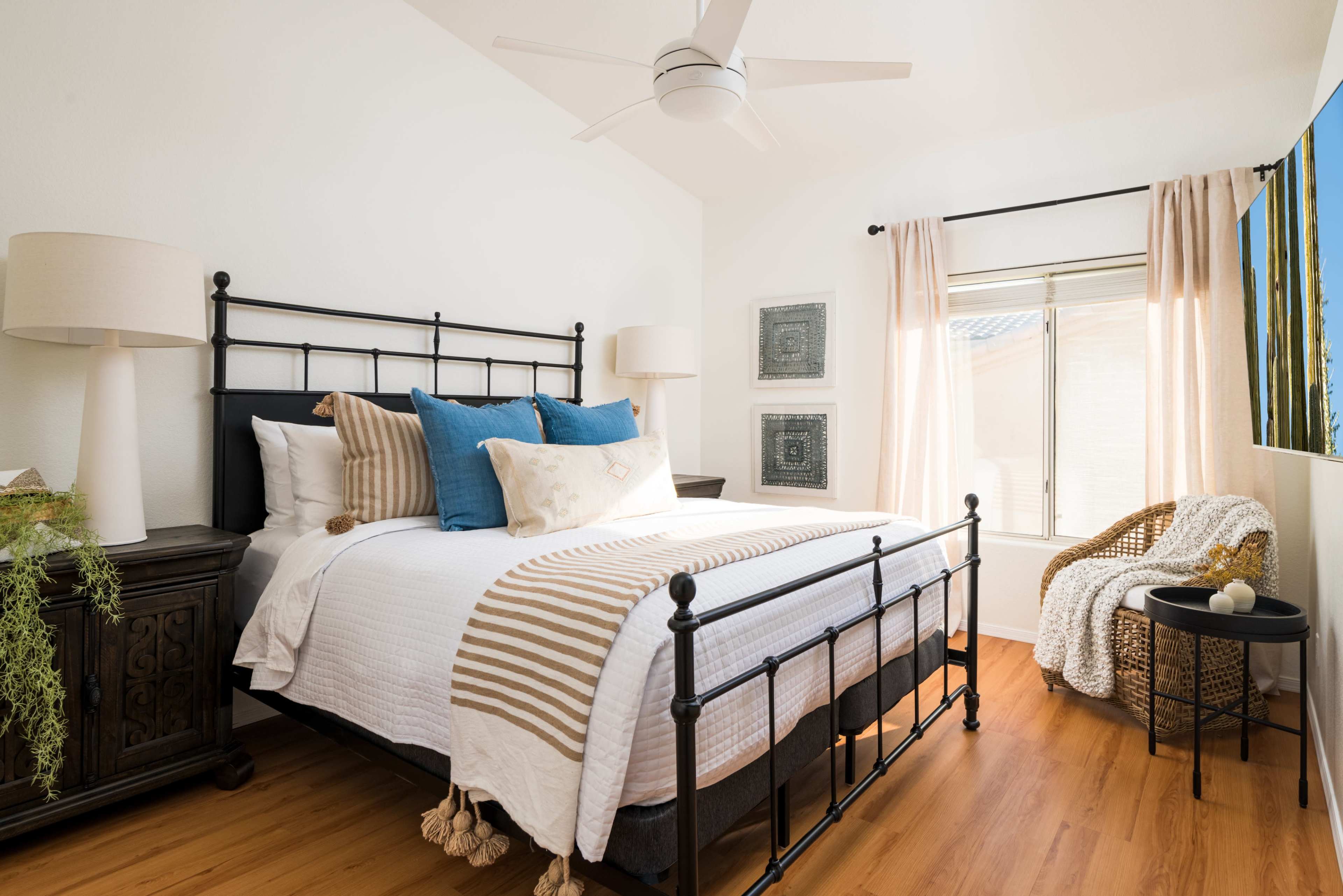A cozy bedroom featuring a metal bed frame with white bedding and blue pillows, a bedside table, a decorative chair, and a large window with natural light.