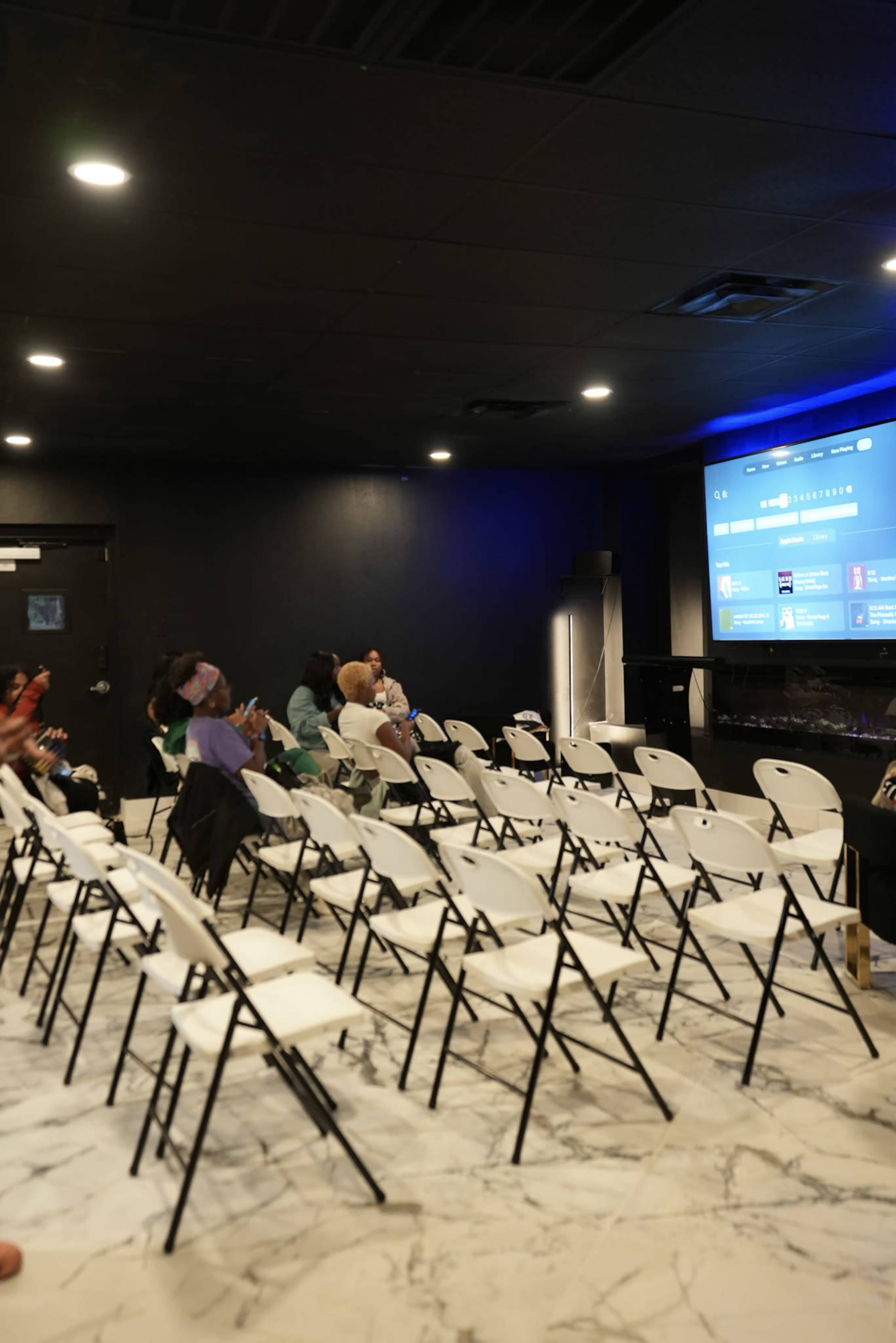 A small audience sits in folding chairs facing a screen in a dimly lit room with marble flooring.