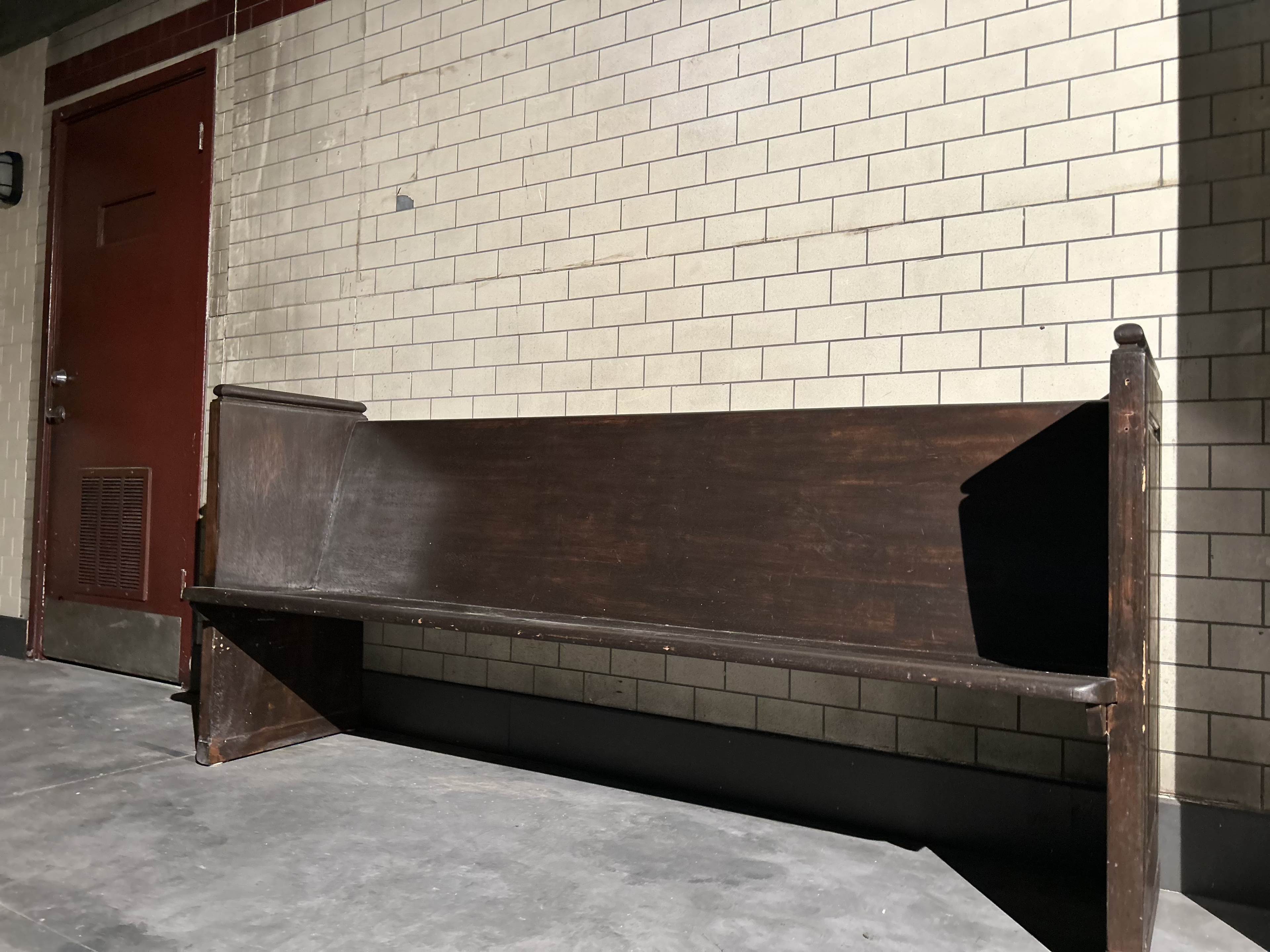 The image shows a weathered wooden bench positioned against a wall with light-colored tiles in a dimly lit corridor.