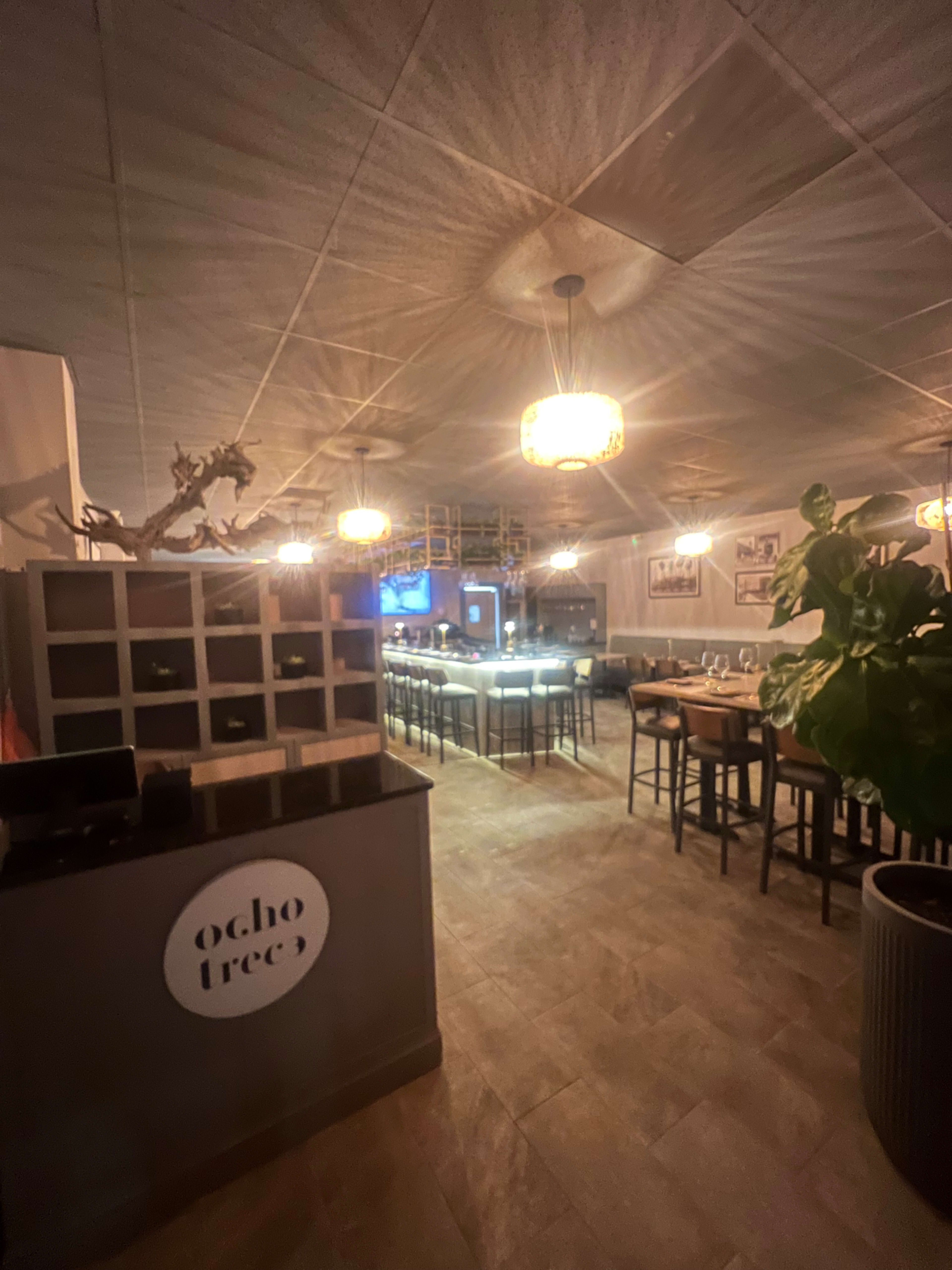 The image shows the interior of a restaurant featuring a bar area, dining tables, and warm lighting.