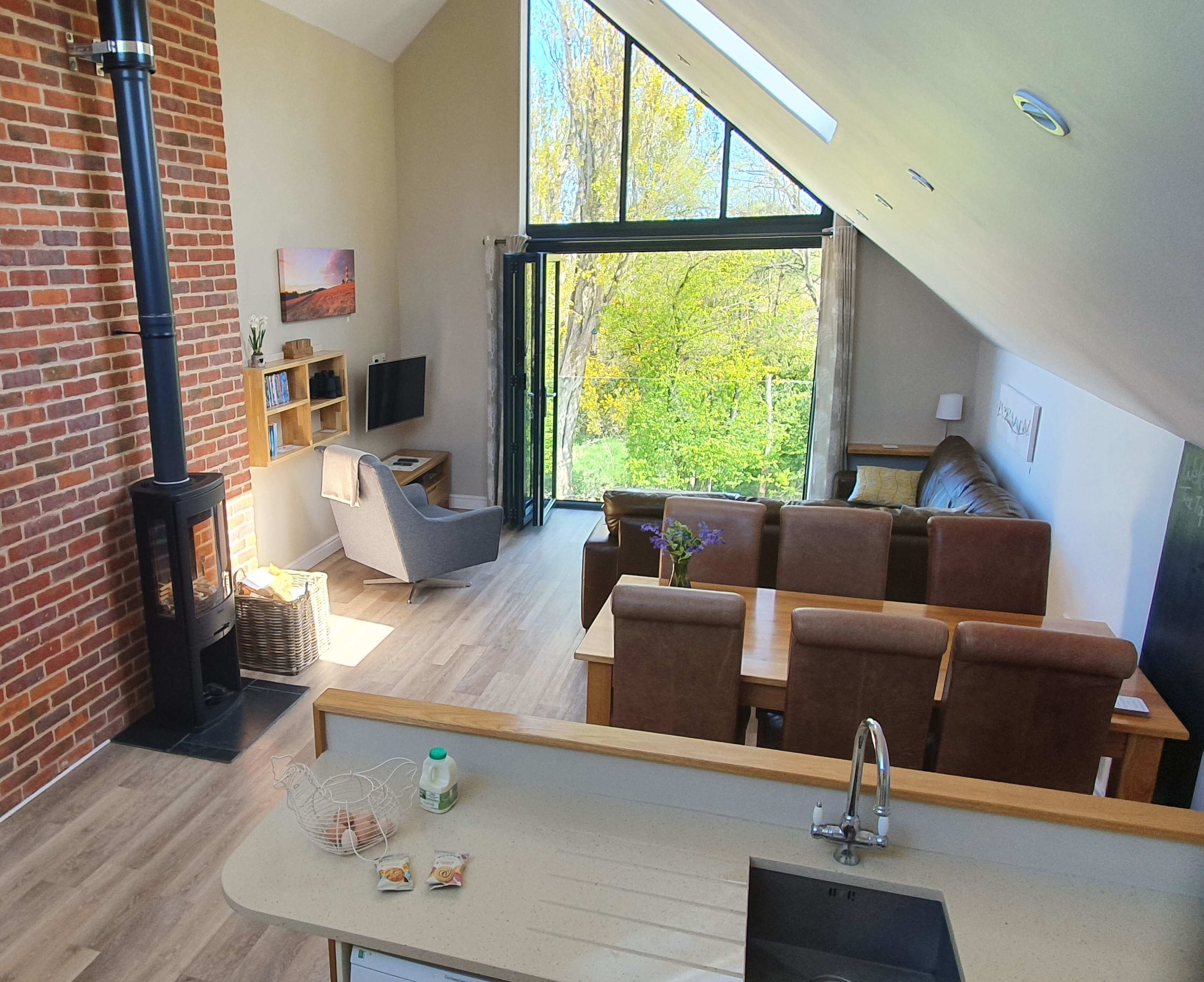 The image shows a modern open-concept living area with a kitchen, dining table, and large windows overlooking a green outdoor space.