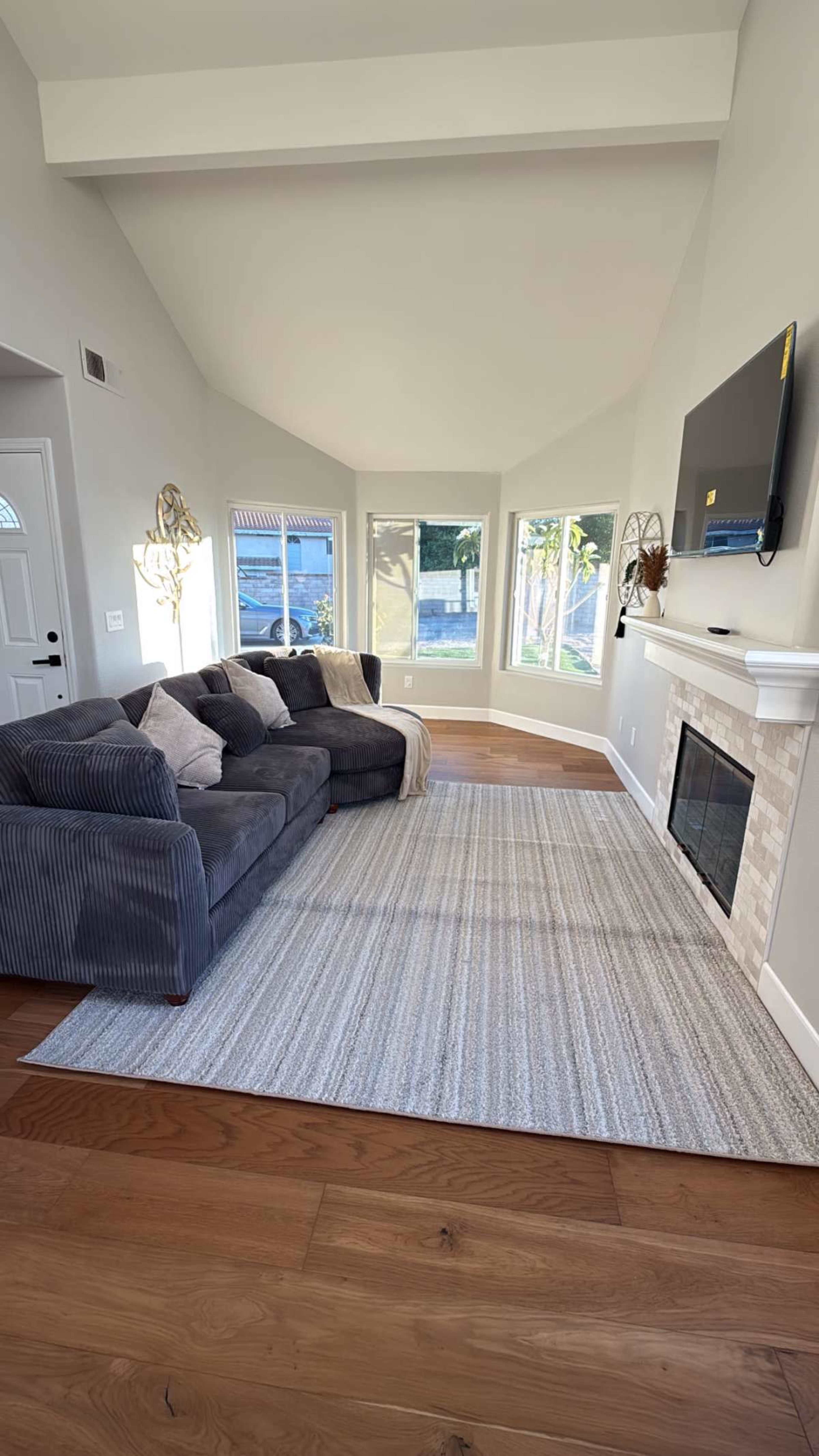 A modern living room features a gray sectional sofa, a flat-screen TV mounted above a stone fireplace, and large windows allowing natural light to enter.