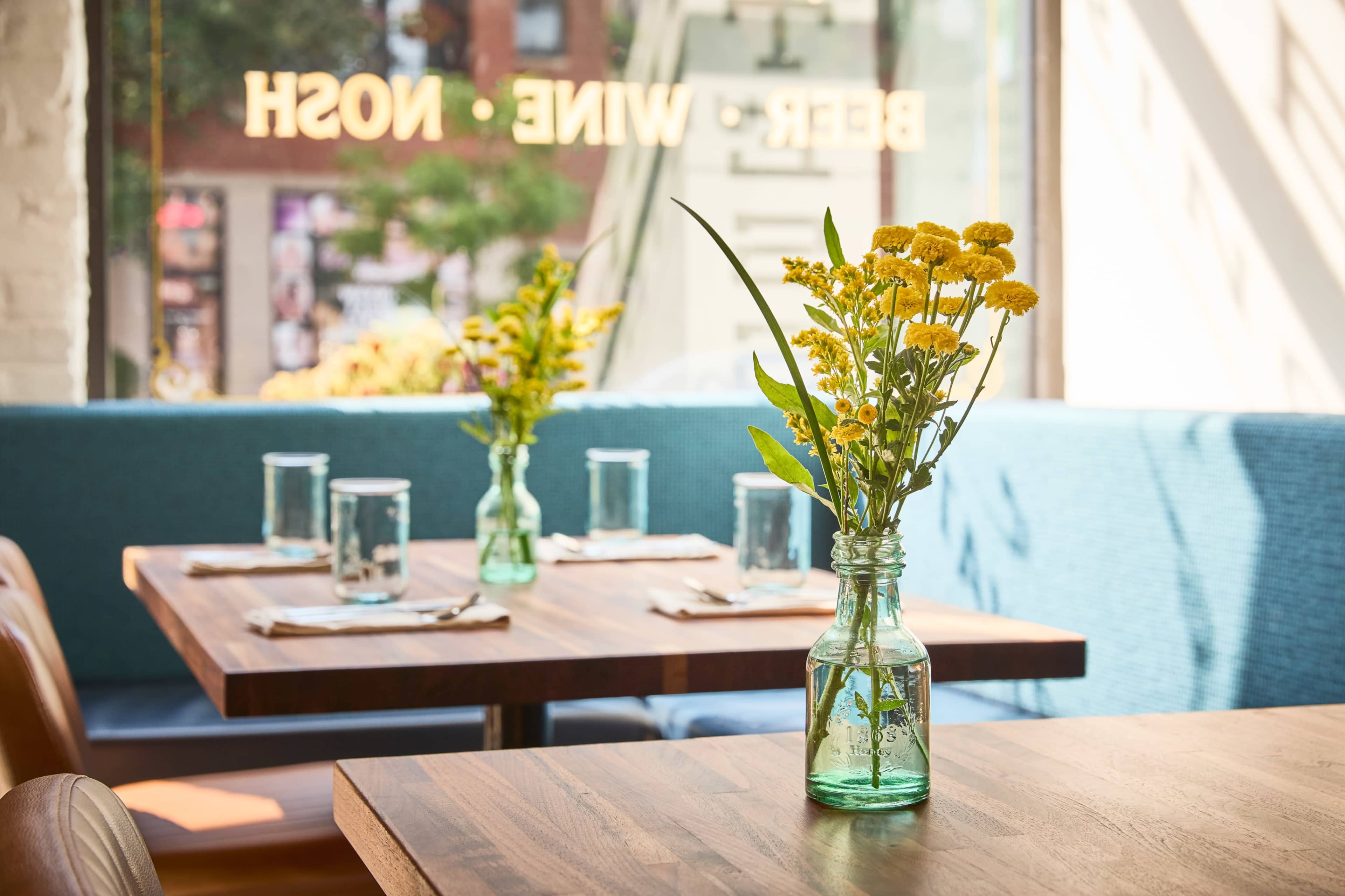 The image shows a bright dining area with wooden tables topped with vases of yellow flowers and glassware, alongside a blue upholstered bench in a restaurant setting.