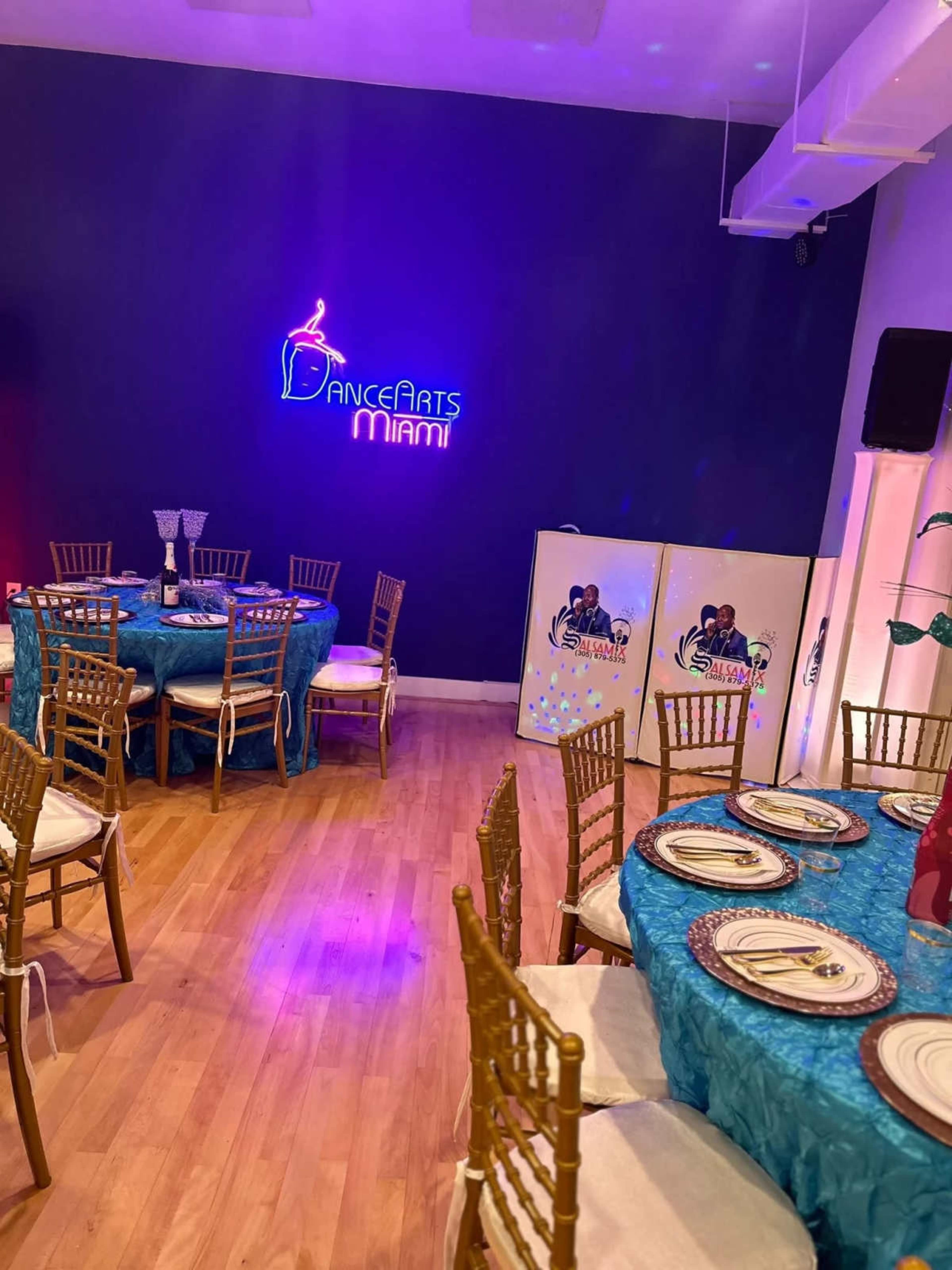 The image shows a decorated event space with tables set for dining, featuring blue tablecloths and gold chairs, along with a neon sign reading "DanceArts Miami" on the wall.