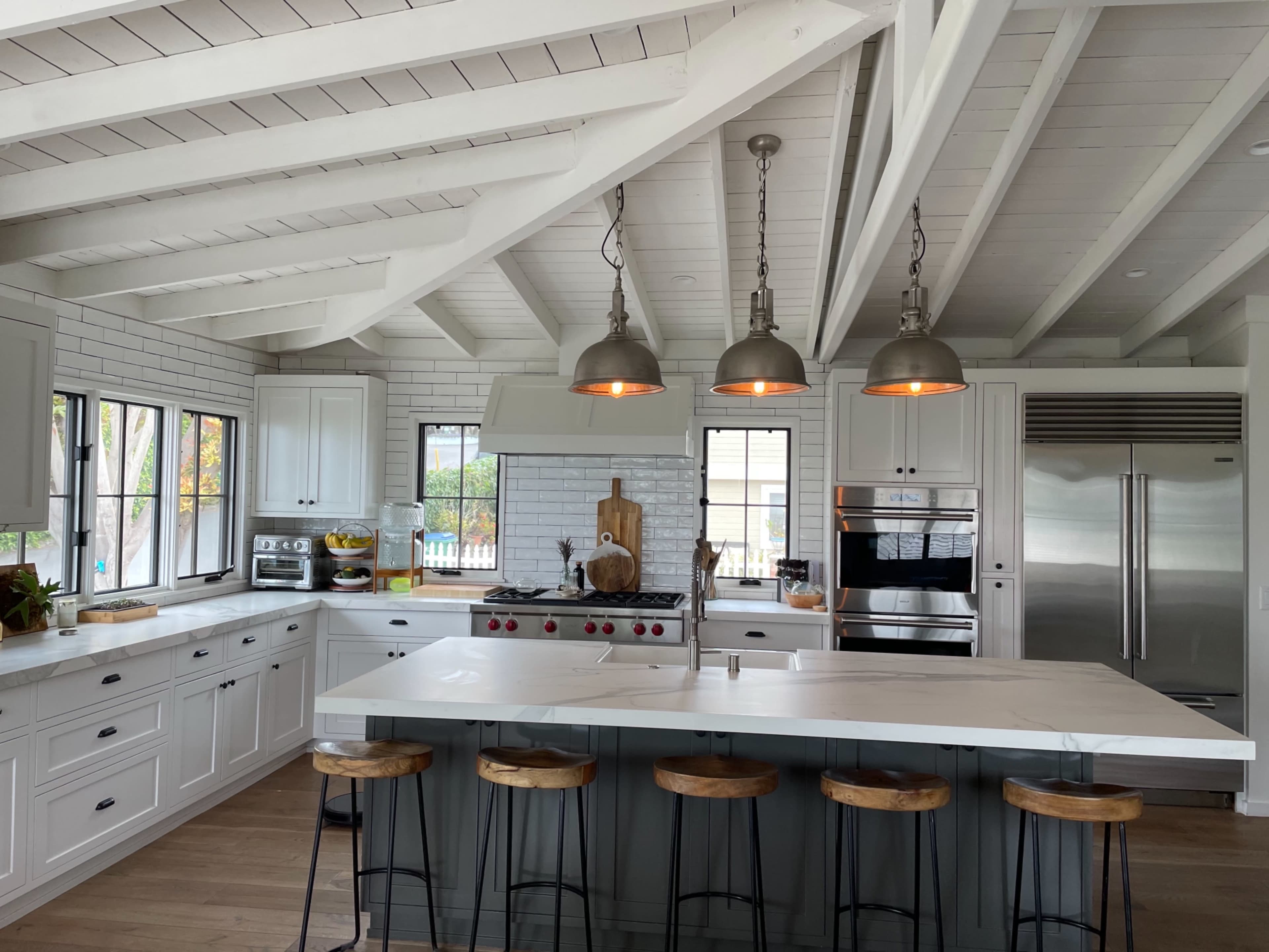 A modern kitchen with a large island, three pendant lights, and stainless steel appliances, featuring white cabinetry and a tiled backsplash.