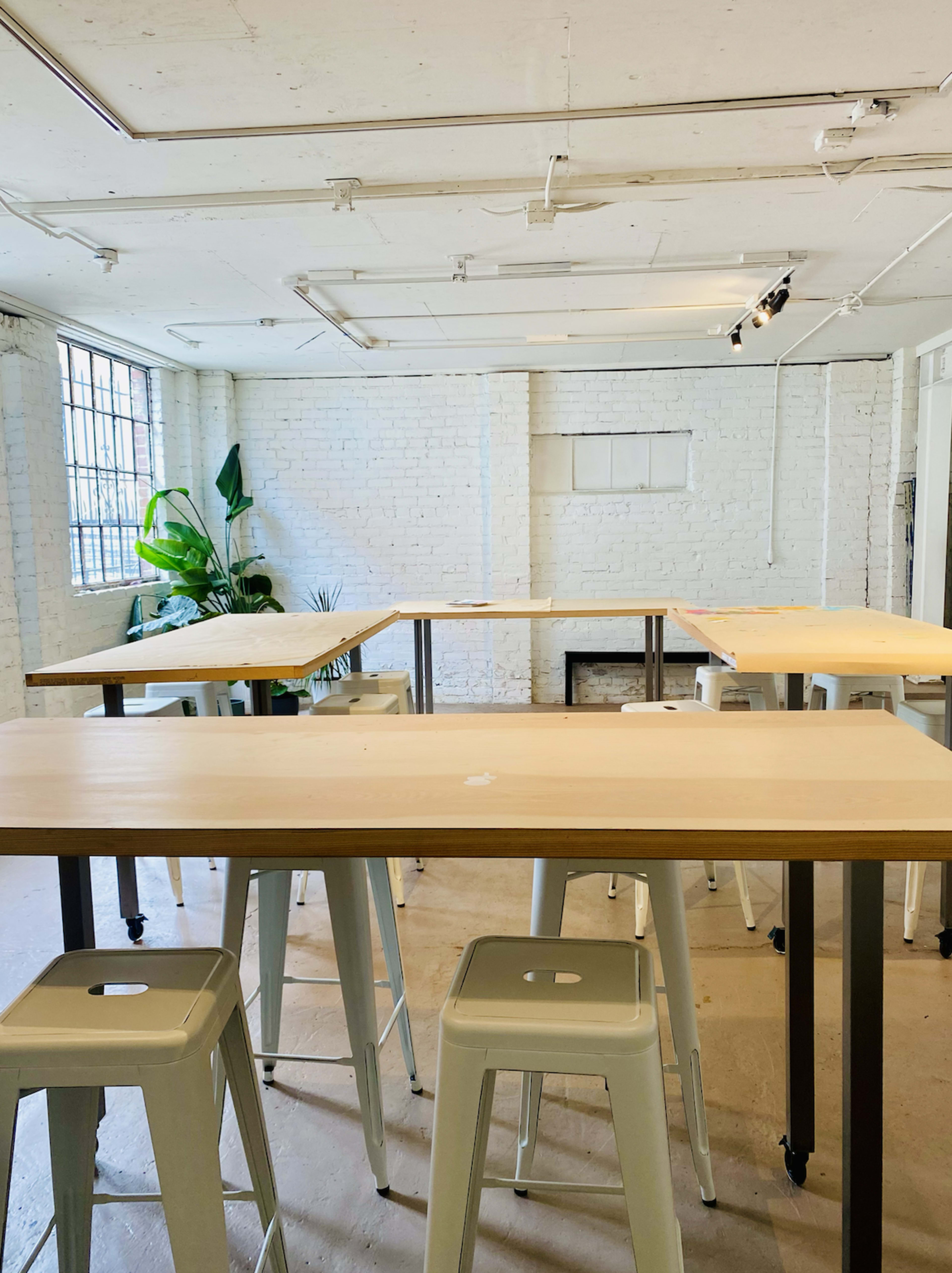 The image shows an open workspace with multiple wooden tables and metal stools, set against a white brick wall and large windows.