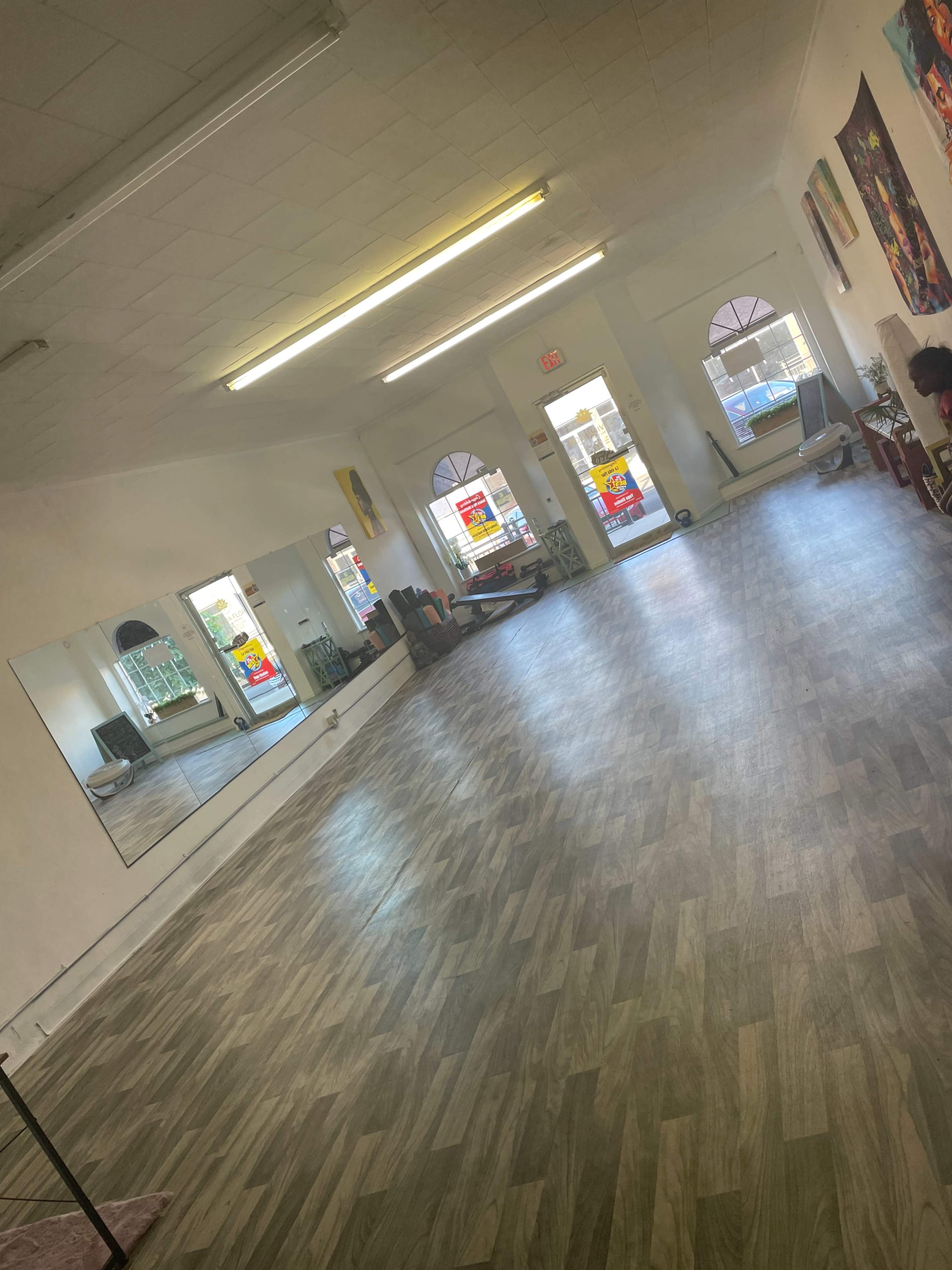 An empty, well-lit room with wood-style flooring, large windows, and a mirror along one wall.