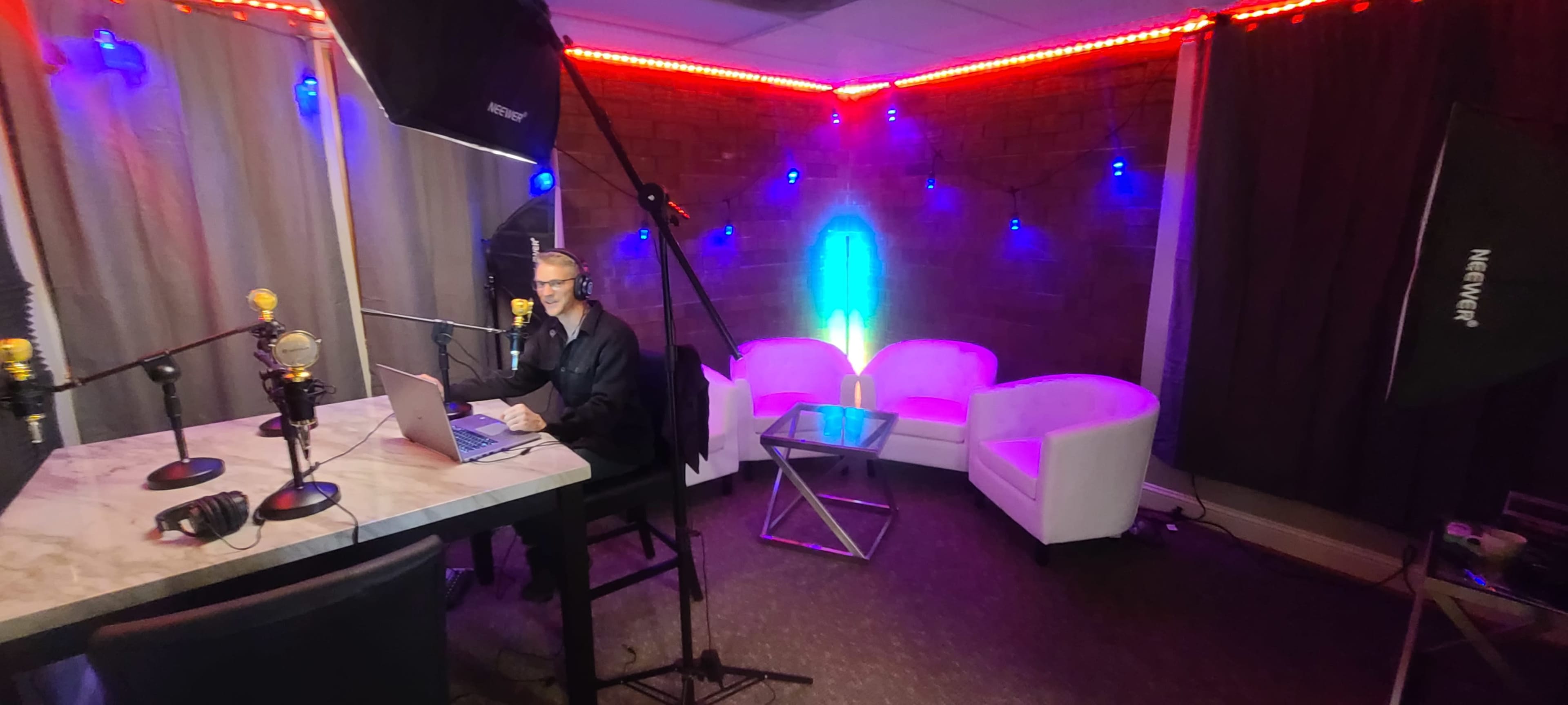 A man sits at a table with a laptop in a brightly lit studio featuring white chairs and multicolored LED lights on the walls.