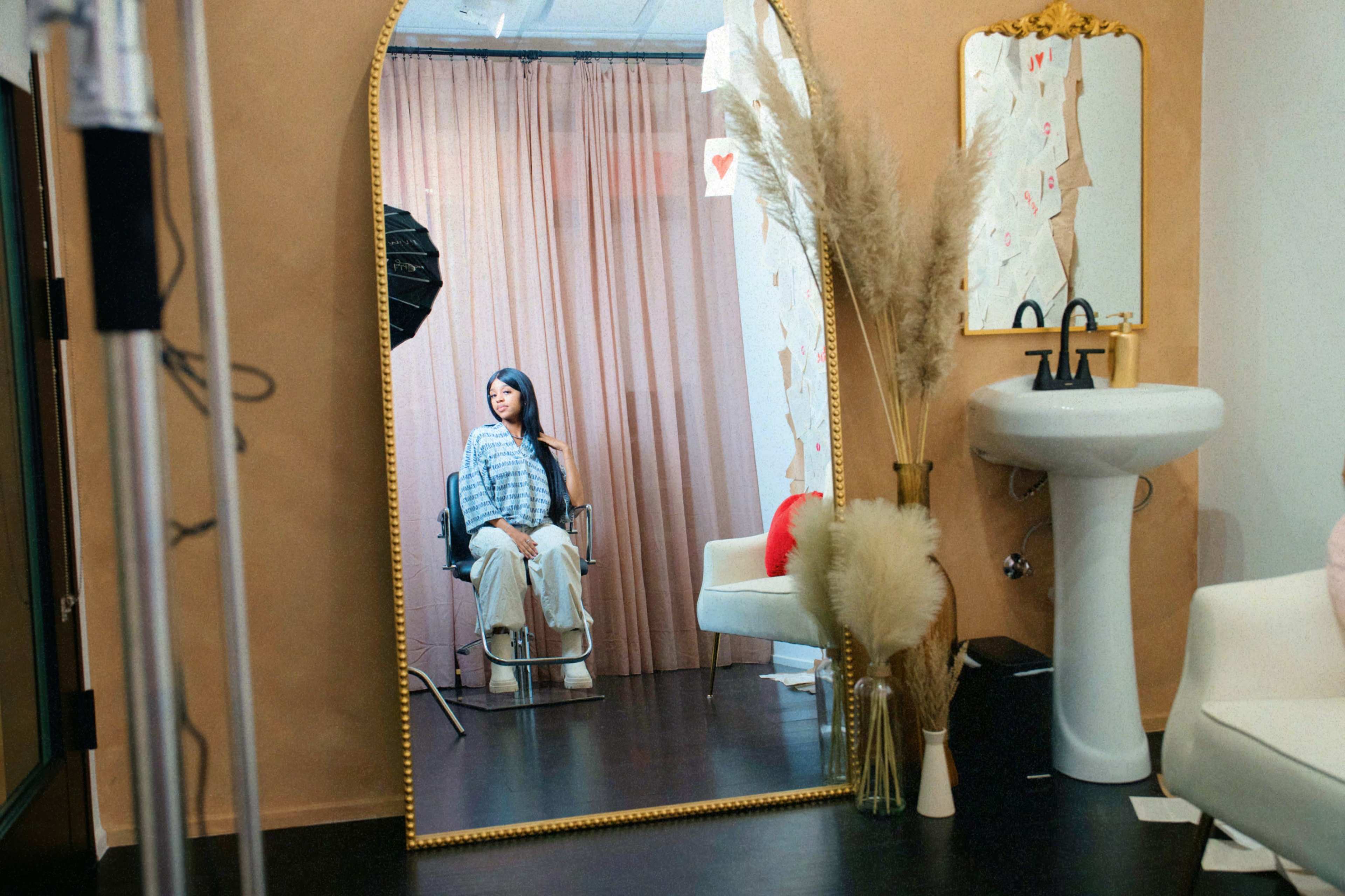 A woman sits in a salon chair in front of a large mirror, surrounded by a backdrop of draped fabric and various decor elements.