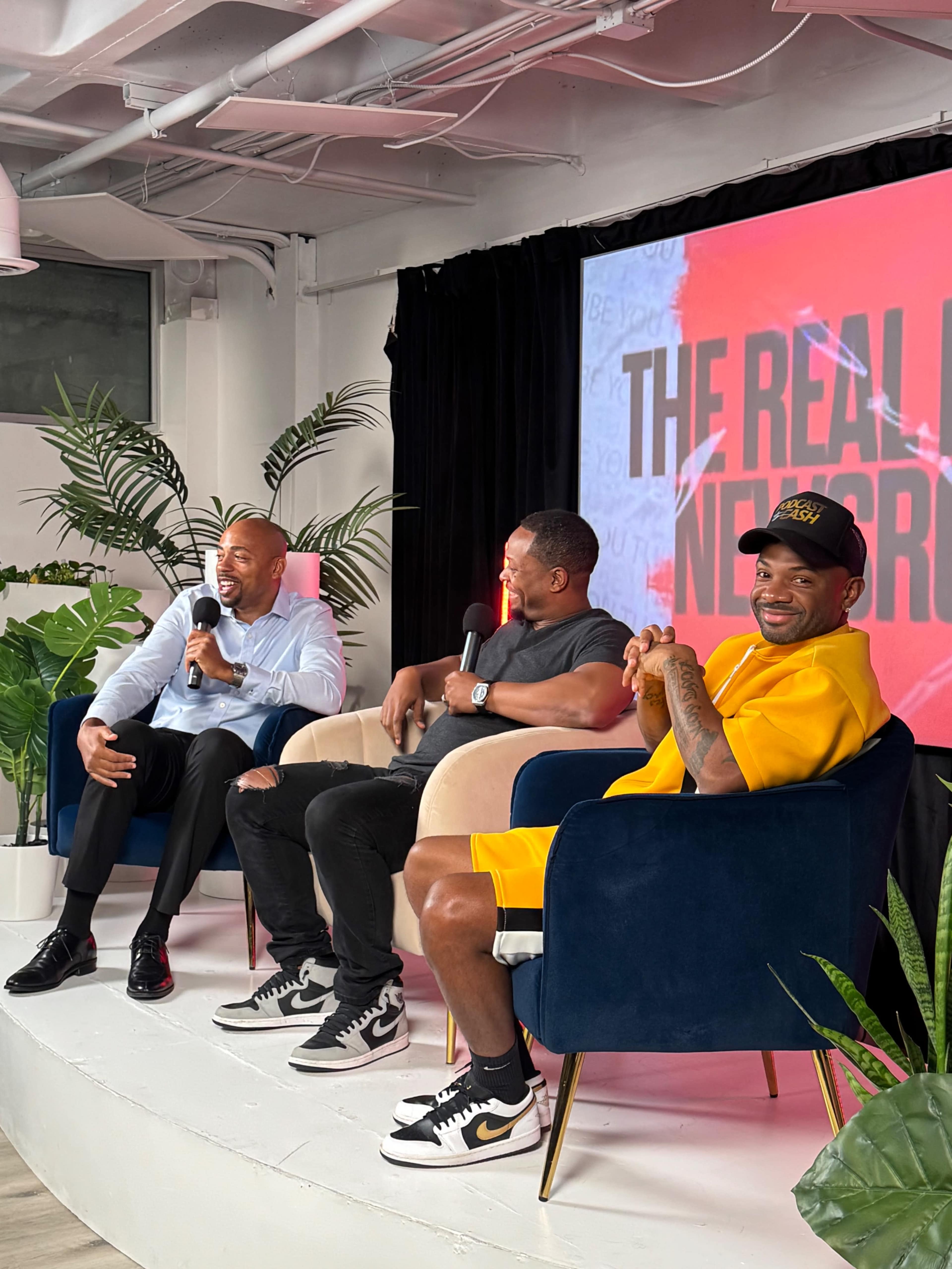 Three men are sitting onstage, engaging in conversation, with a large screen behind them displaying the text "THE REAL NETWORK."