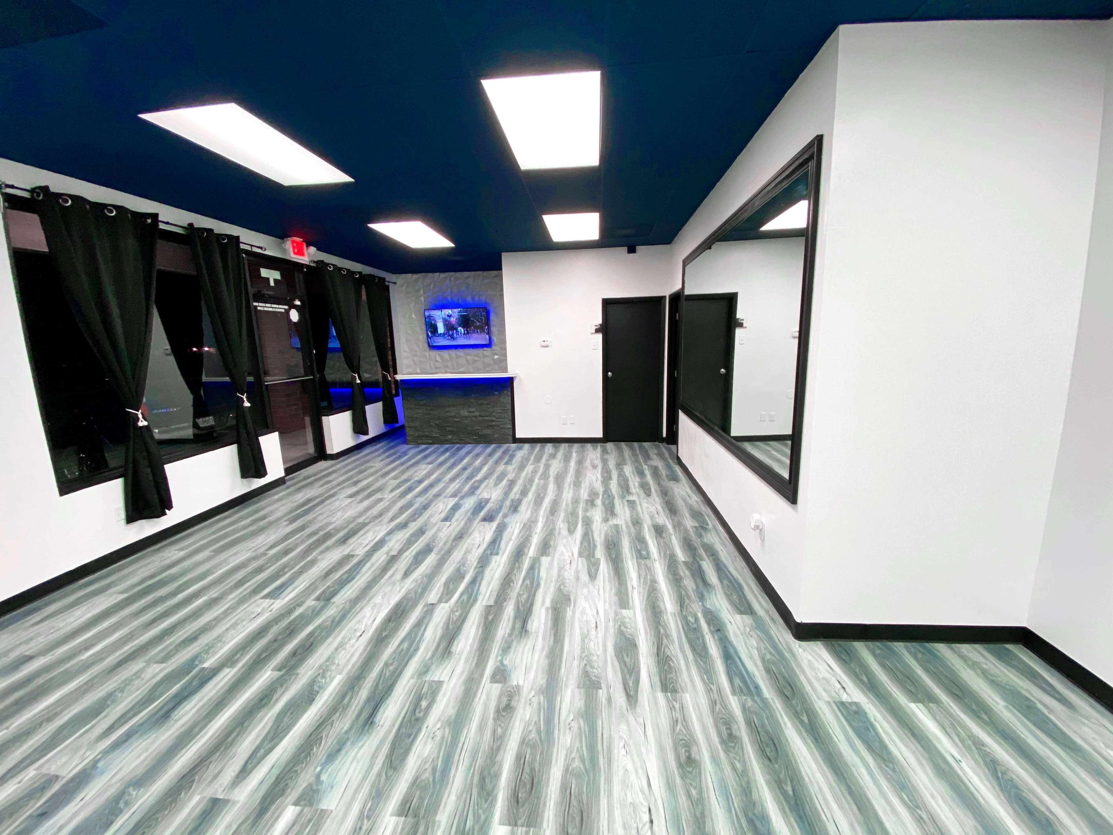 A spacious room with a dark blue ceiling, modern lighting, and a light gray wooden floor, featuring large mirrors and black curtains along the windows.