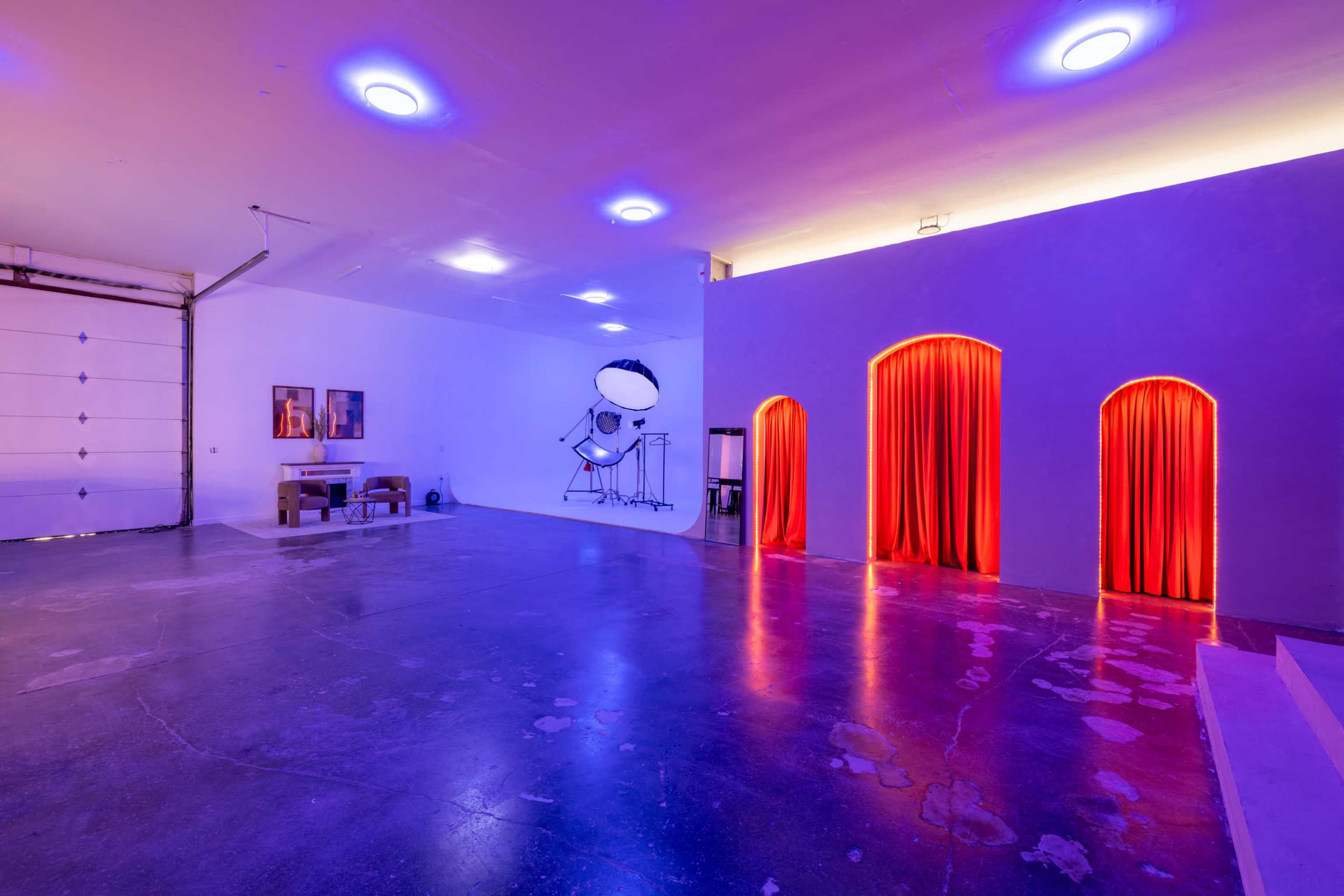The image shows a spacious, modern interior with a polished concrete floor illuminated by purple lighting, featuring a large garage door, a seating area, and three arched openings with red curtains.