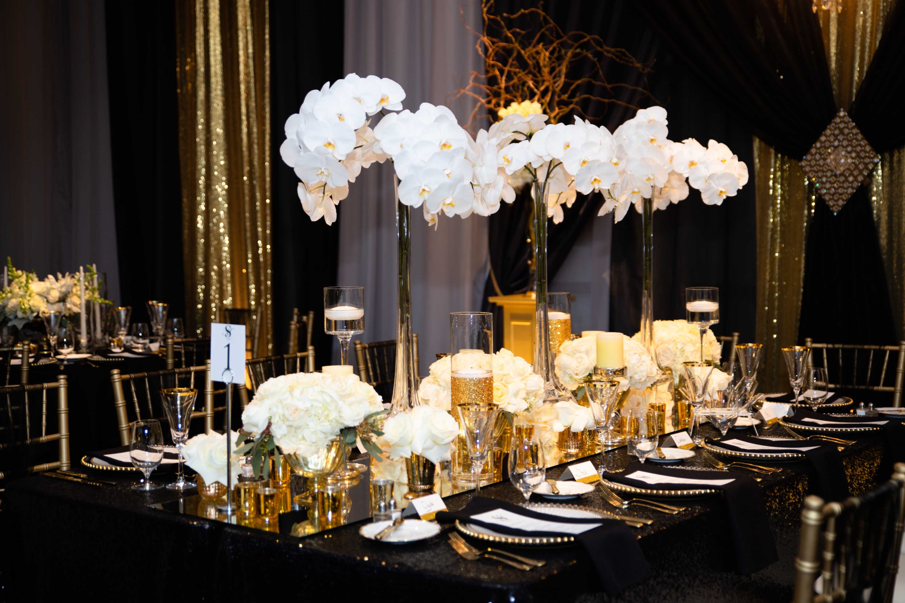The table is elegantly set with tall white orchid centerpieces, gold accents, and black tableware against a backdrop of shimmering gold and black drapery.