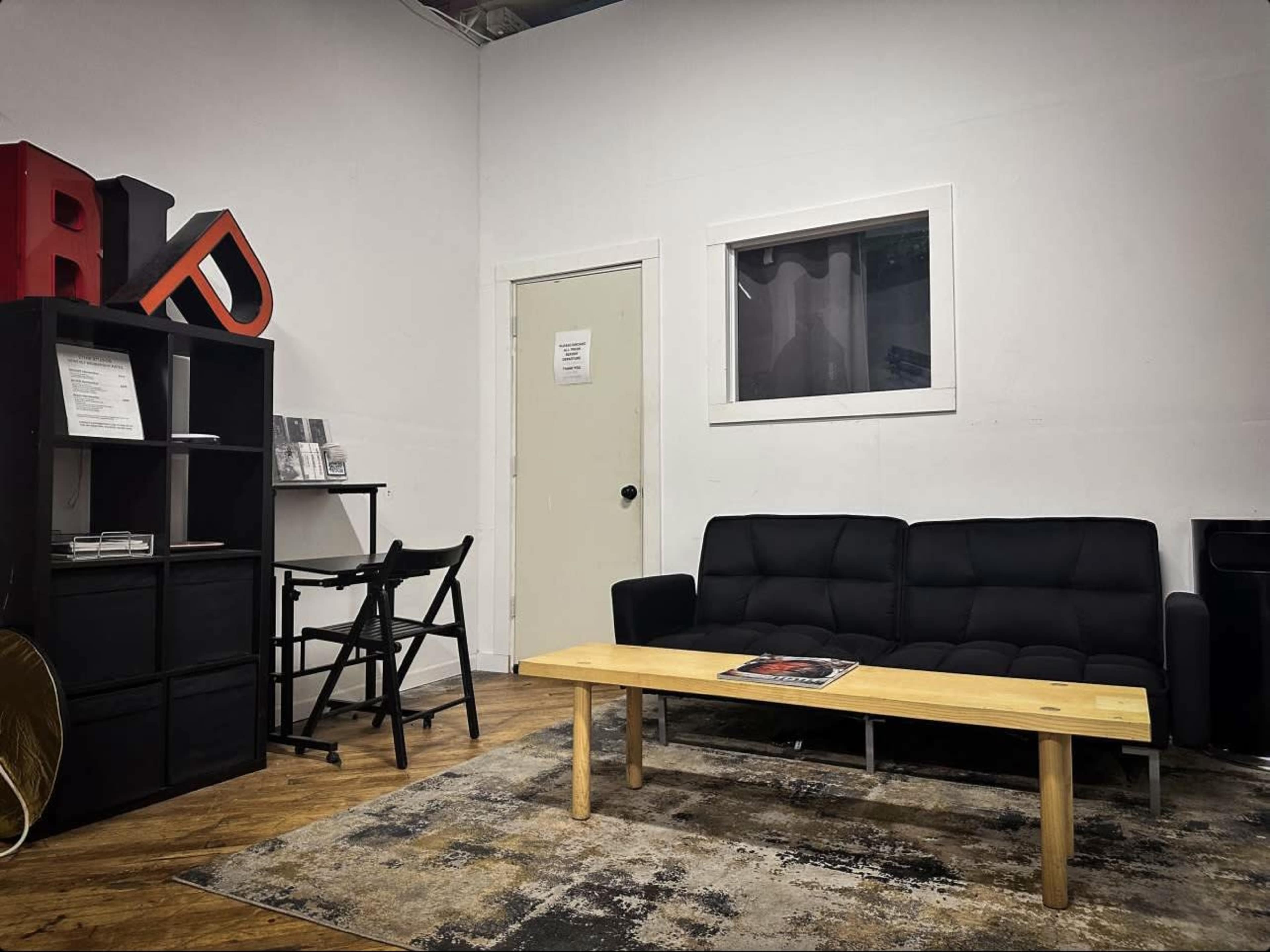 The image depicts a minimalist studio space featuring a black sofa, a wooden coffee table, a bookshelf, and a folding chair against a white wall.