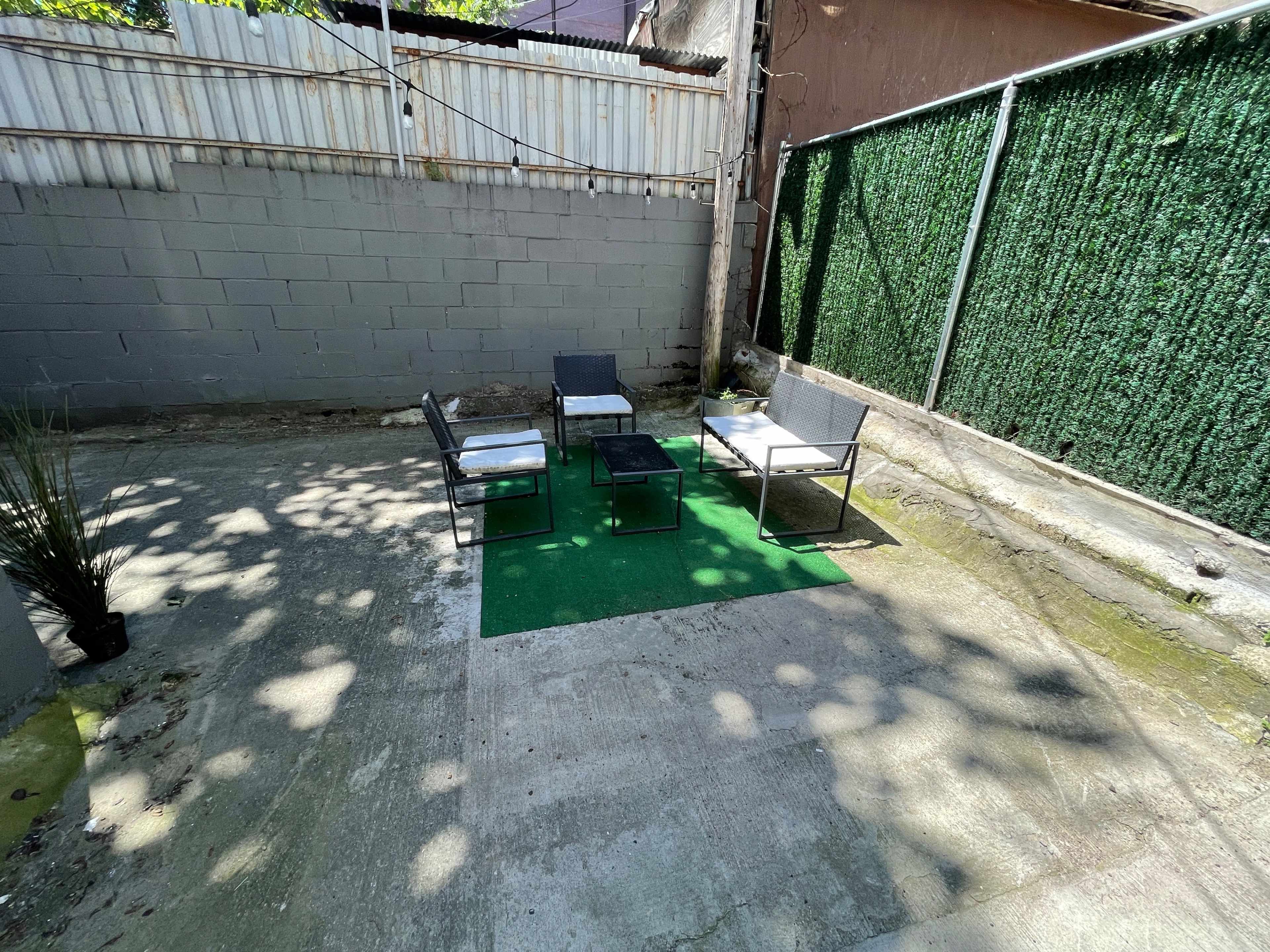 A small, concrete outdoor area featuring four chairs and a table arranged on a green mat, with a wall covered in green foliage in the background.
