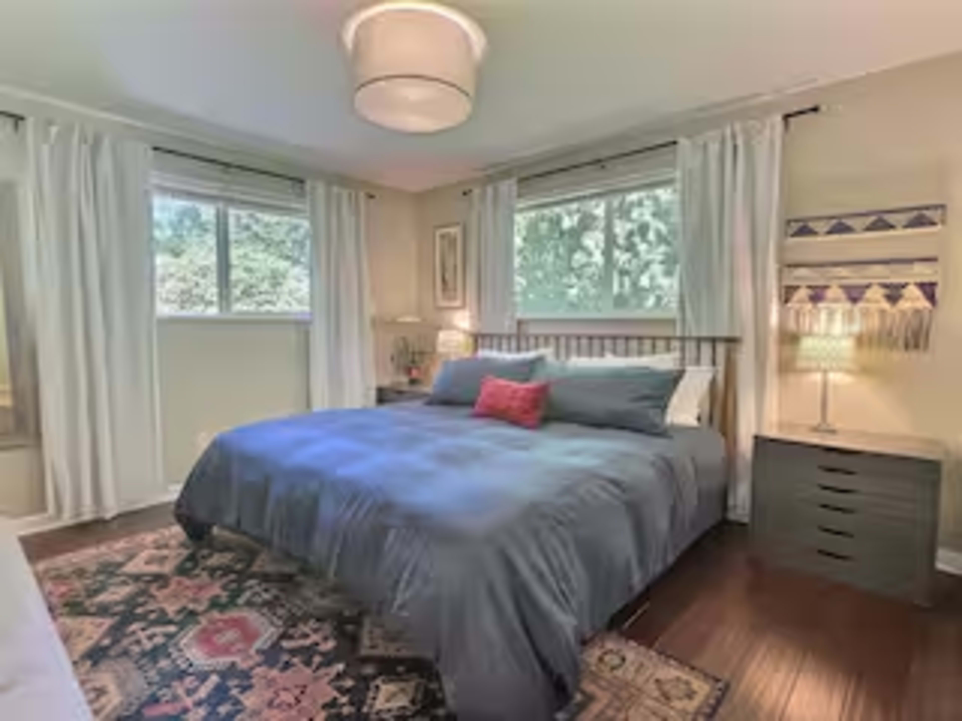 The image shows a well-lit bedroom featuring a queen-sized bed with blue bedding, two windows with sheer white curtains, and a nightstand beside the bed.