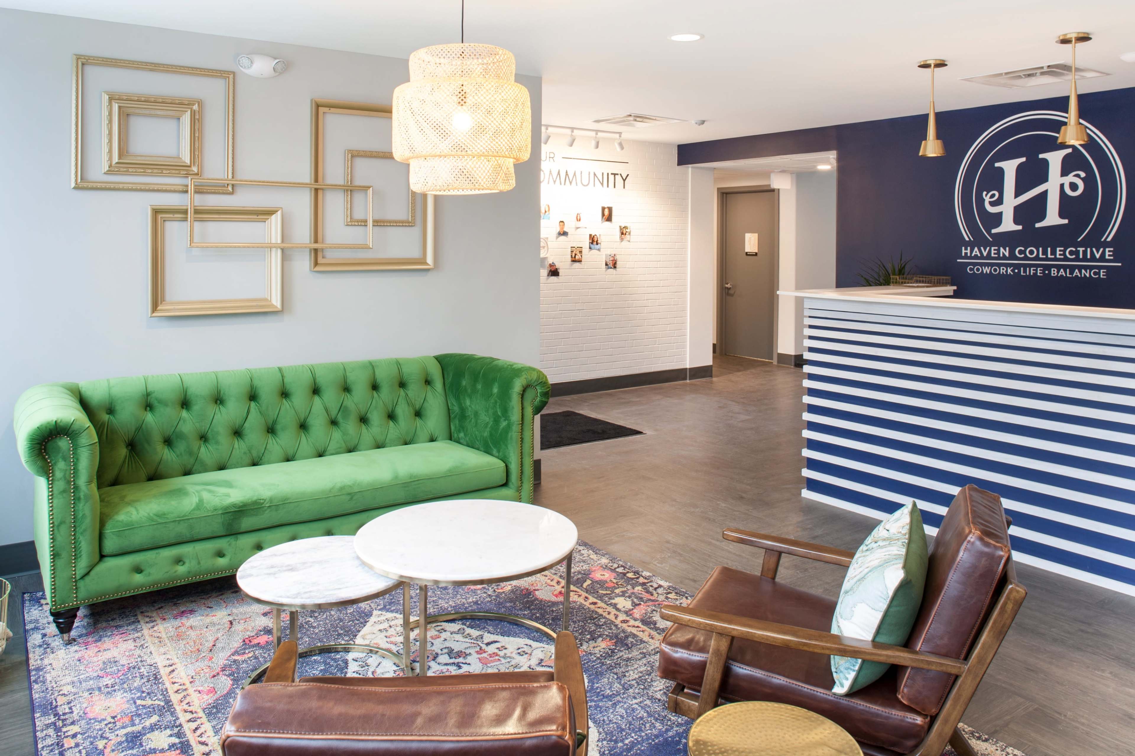 A modern reception area featuring a green tufted sofa, two brown leather chairs, and a striped front desk with decorative wall frames and a logo on display.