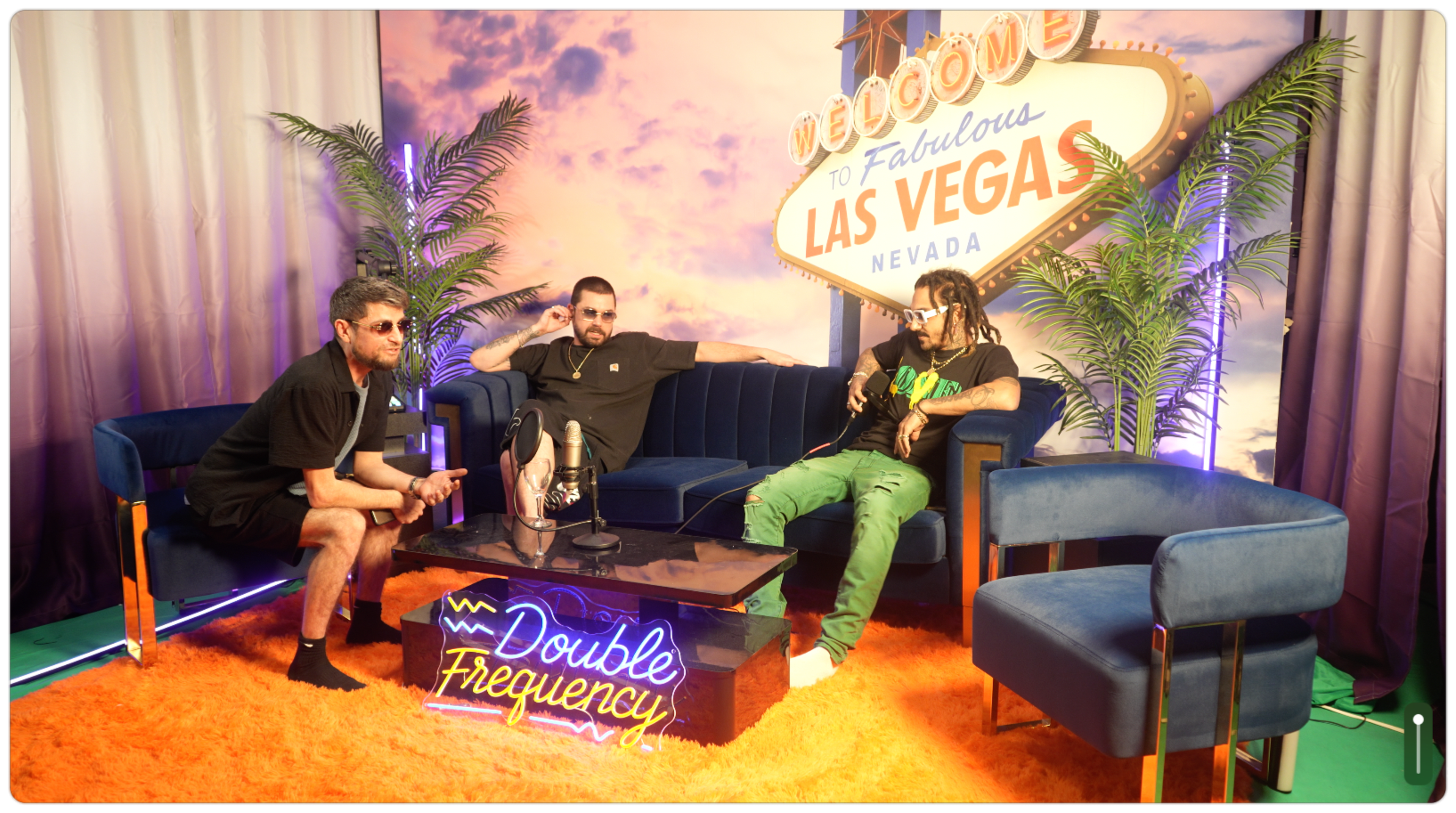 Three men are seated on blue couches in front of a colorful backdrop featuring "Welcome to Fabulous Las Vegas" signage, with a neon "Double Frequency" sign illuminated on a coffee table.