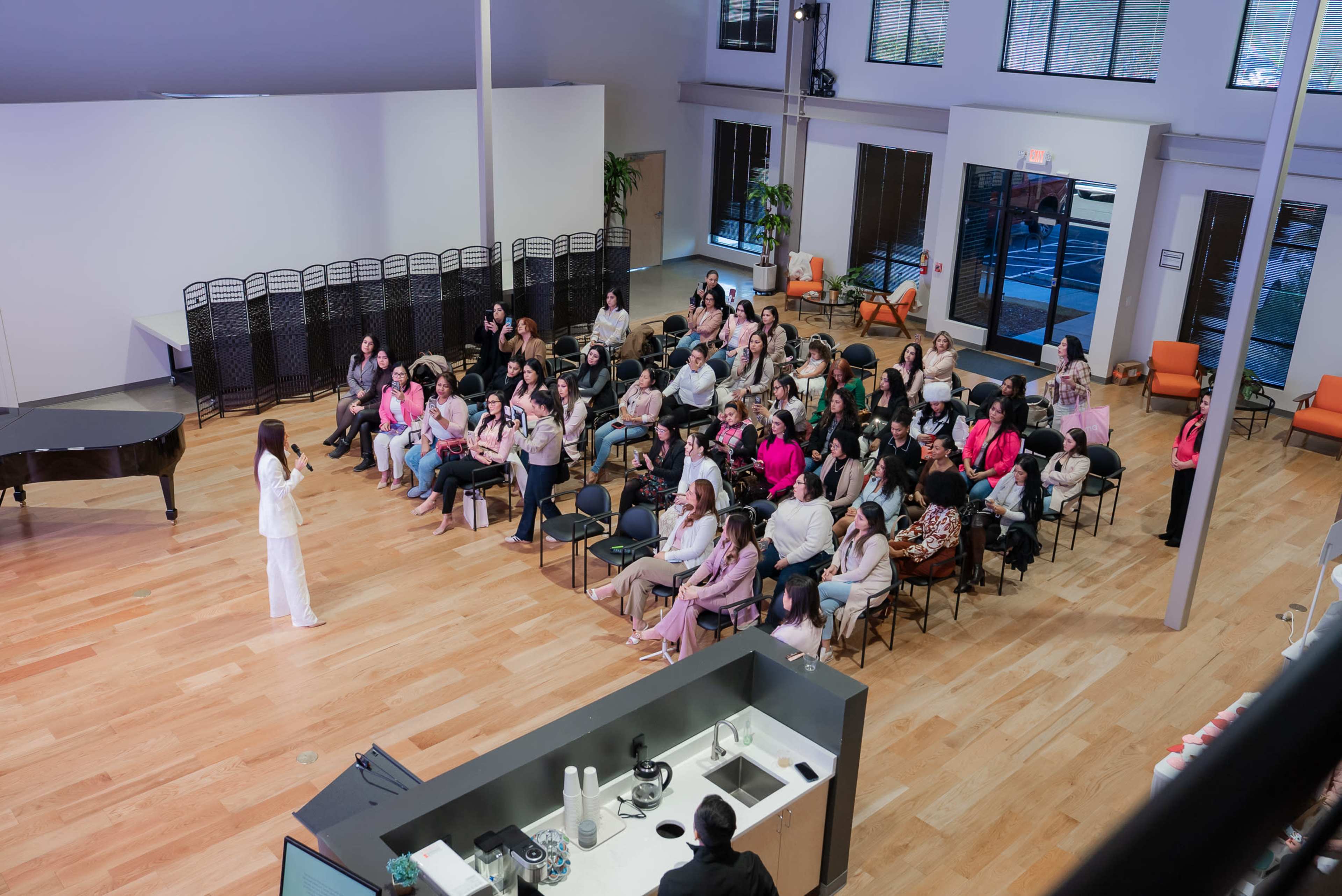 A speaker addresses an audience seated in a spacious, bright room with large windows and a piano in the background.