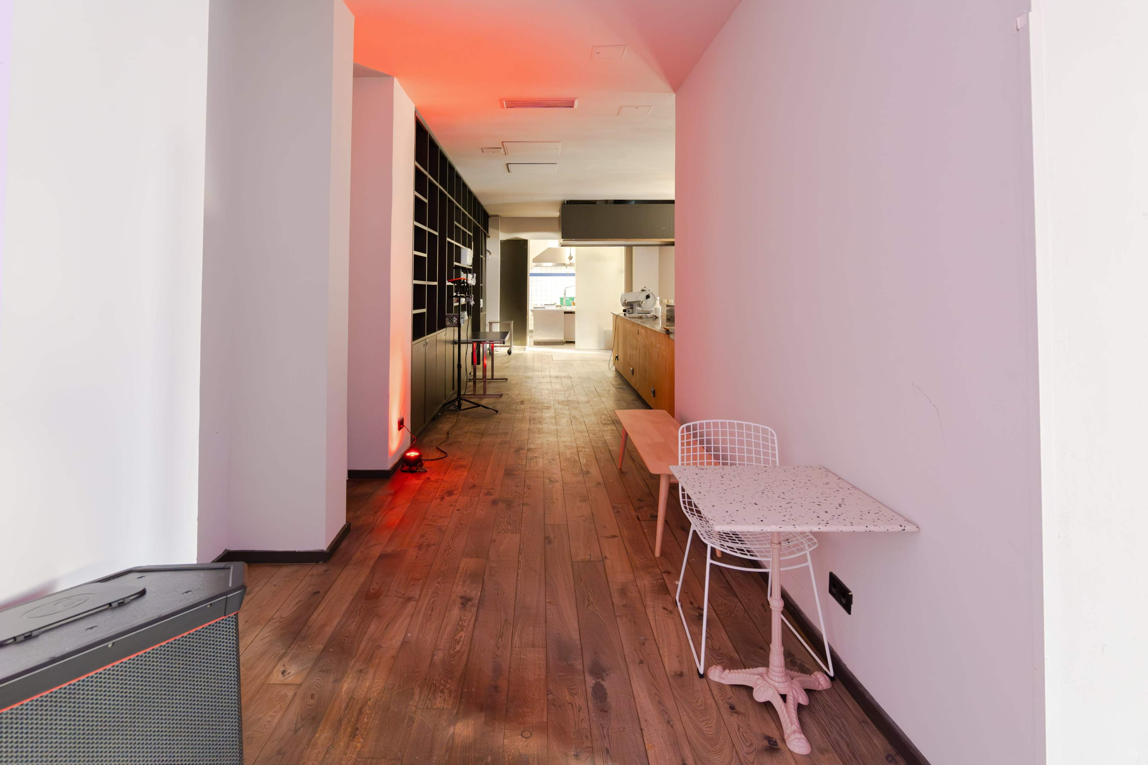 A narrow corridor leading to a café area, with wooden flooring, a small table, and shelves on one side.