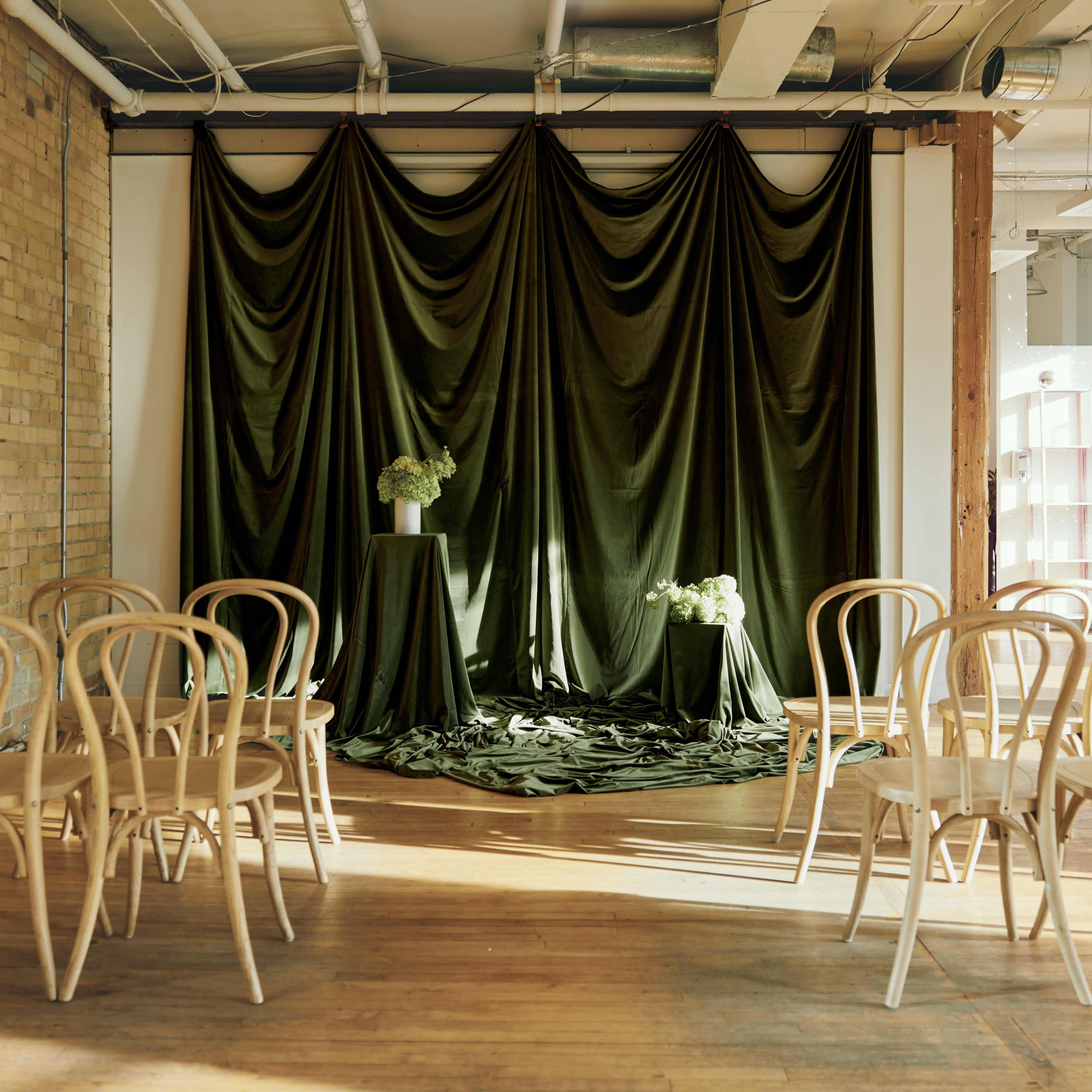The scene features a room with a wooden floor, adorned with draped olive green fabric and a small pedestal holding flowers, surrounded by empty wooden chairs arranged in rows.