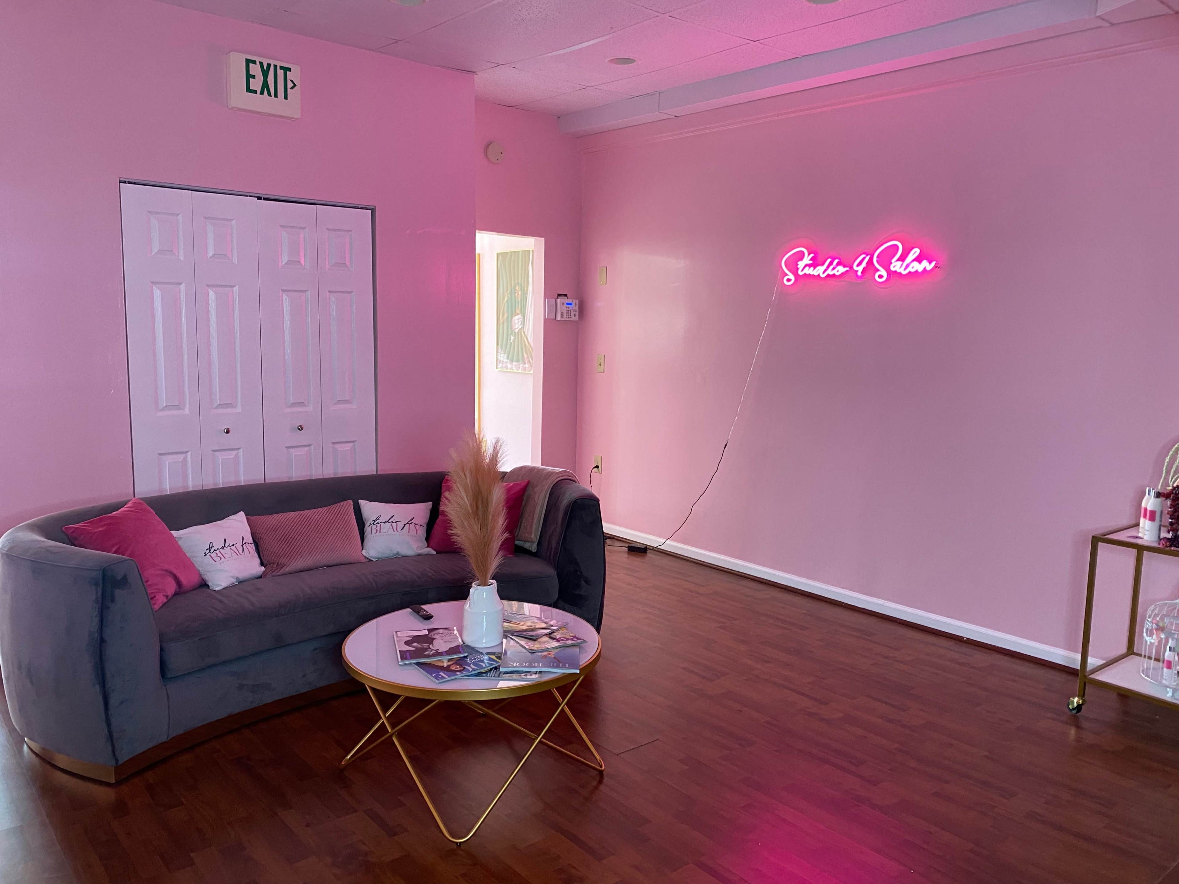 A pink-walled room features a gray couch with decorative pillows, a round coffee table, and a neon sign that reads "Studio & Salon."