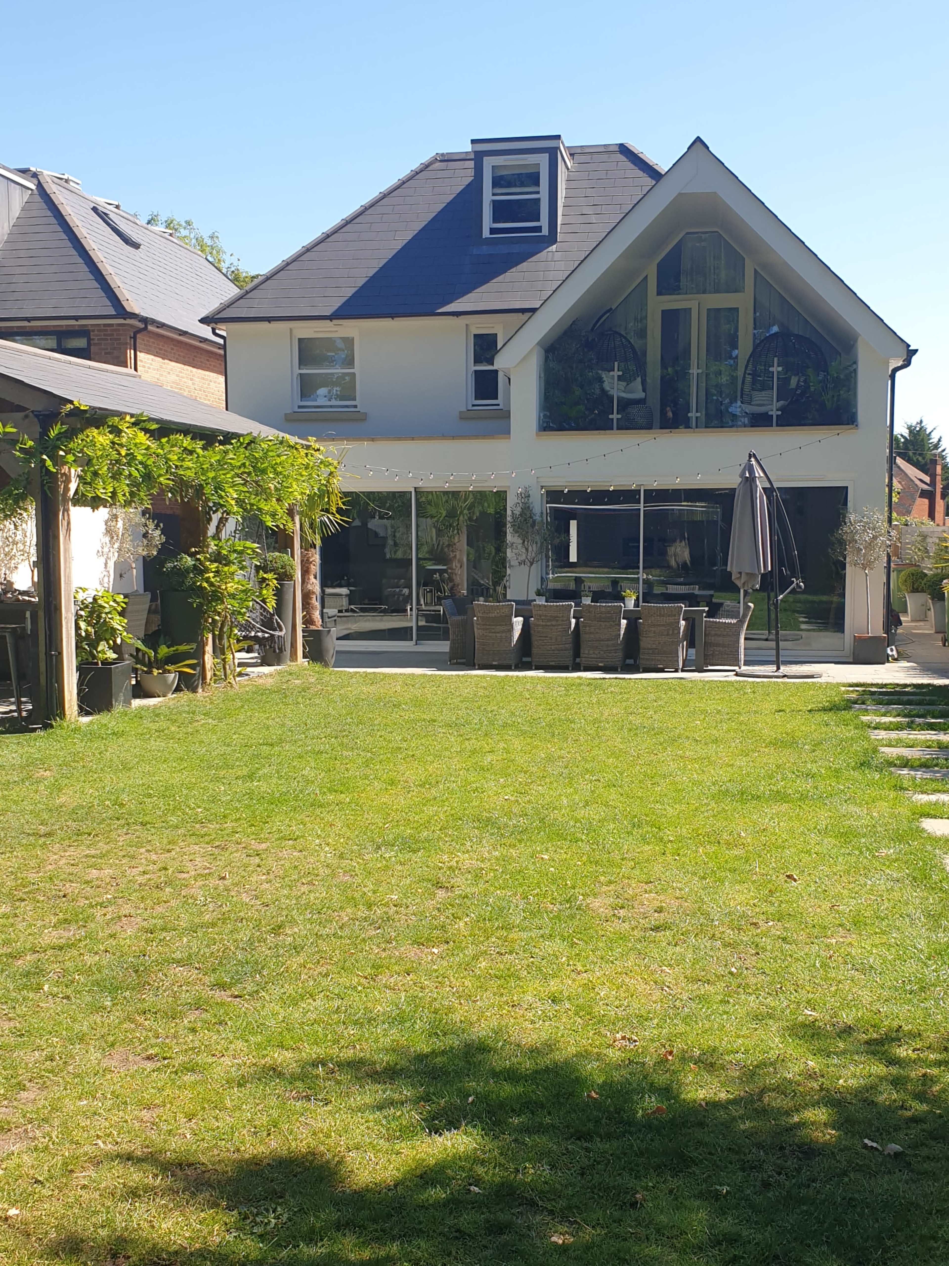 A modern two-story house with a large glass patio overlooks a well-maintained green lawn and patio seating area.